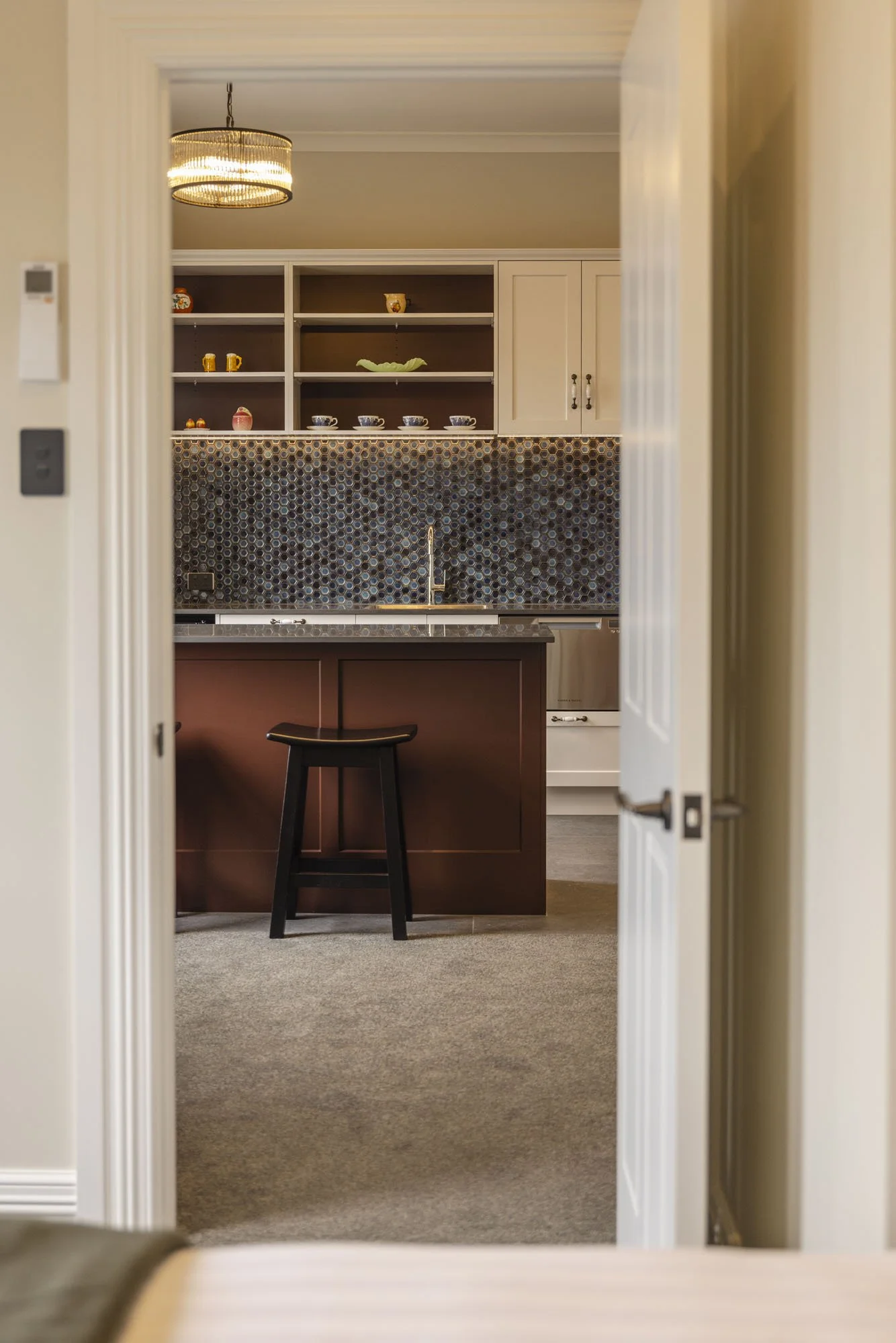 View through doorway into a kitchen with open shelving, dark mosaic tile backsplash, and a brown kitchen island with a black stool.