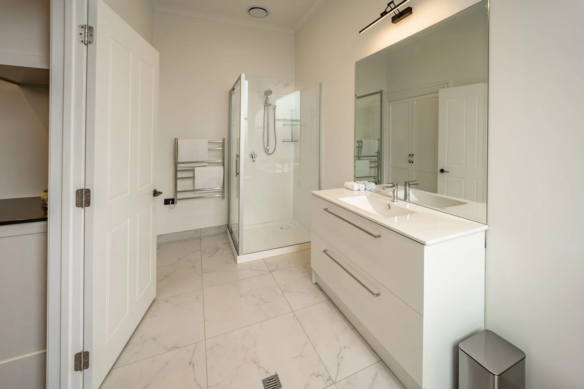 Modern bathroom with white walls, a glass shower enclosure, a white vanity with a large mirror, and marble floor tiles.
