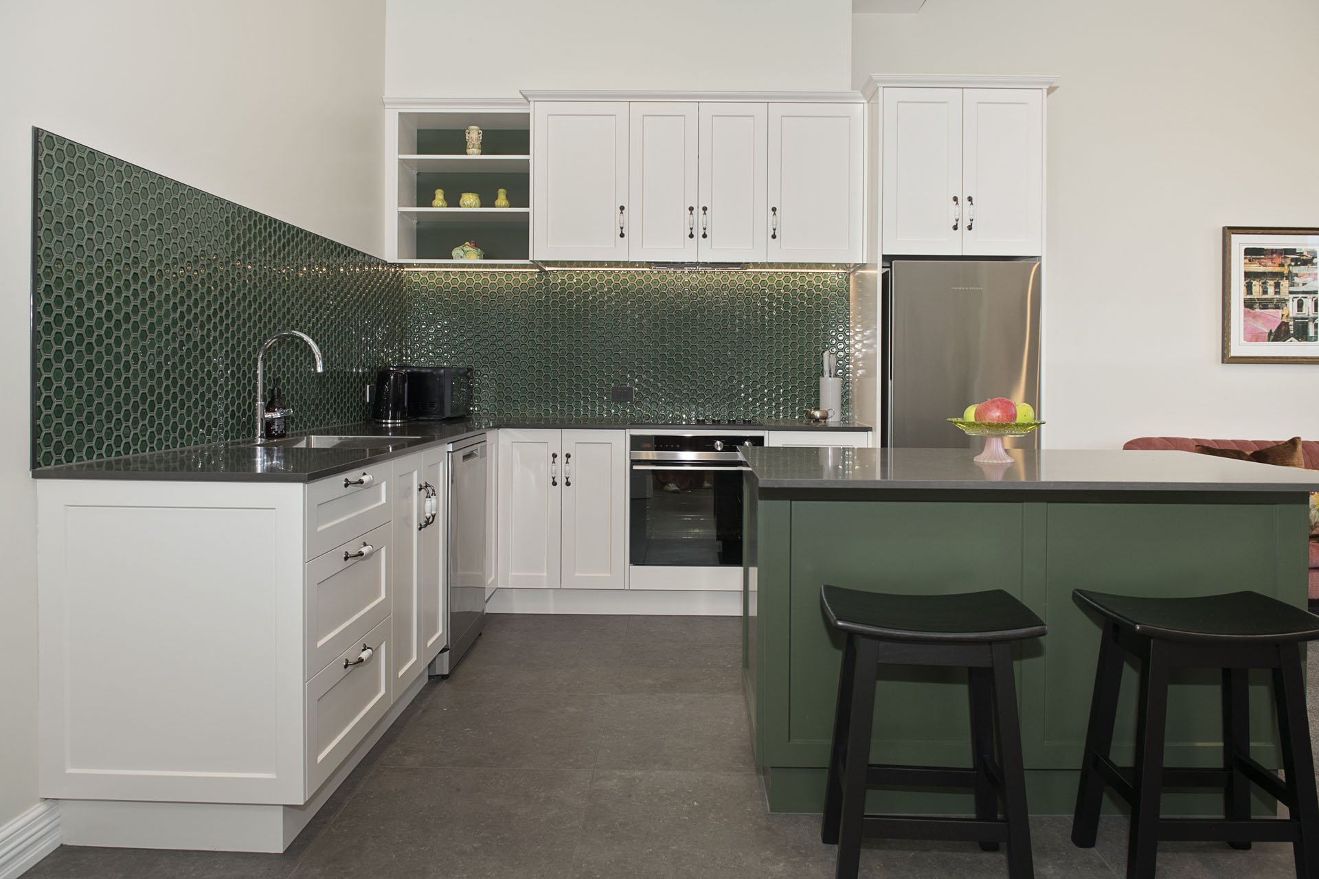 Modern kitchen with white cabinets, green hexagonal tile backsplash, black countertop, and a kitchen island with black stools, featuring a bowl of apples.