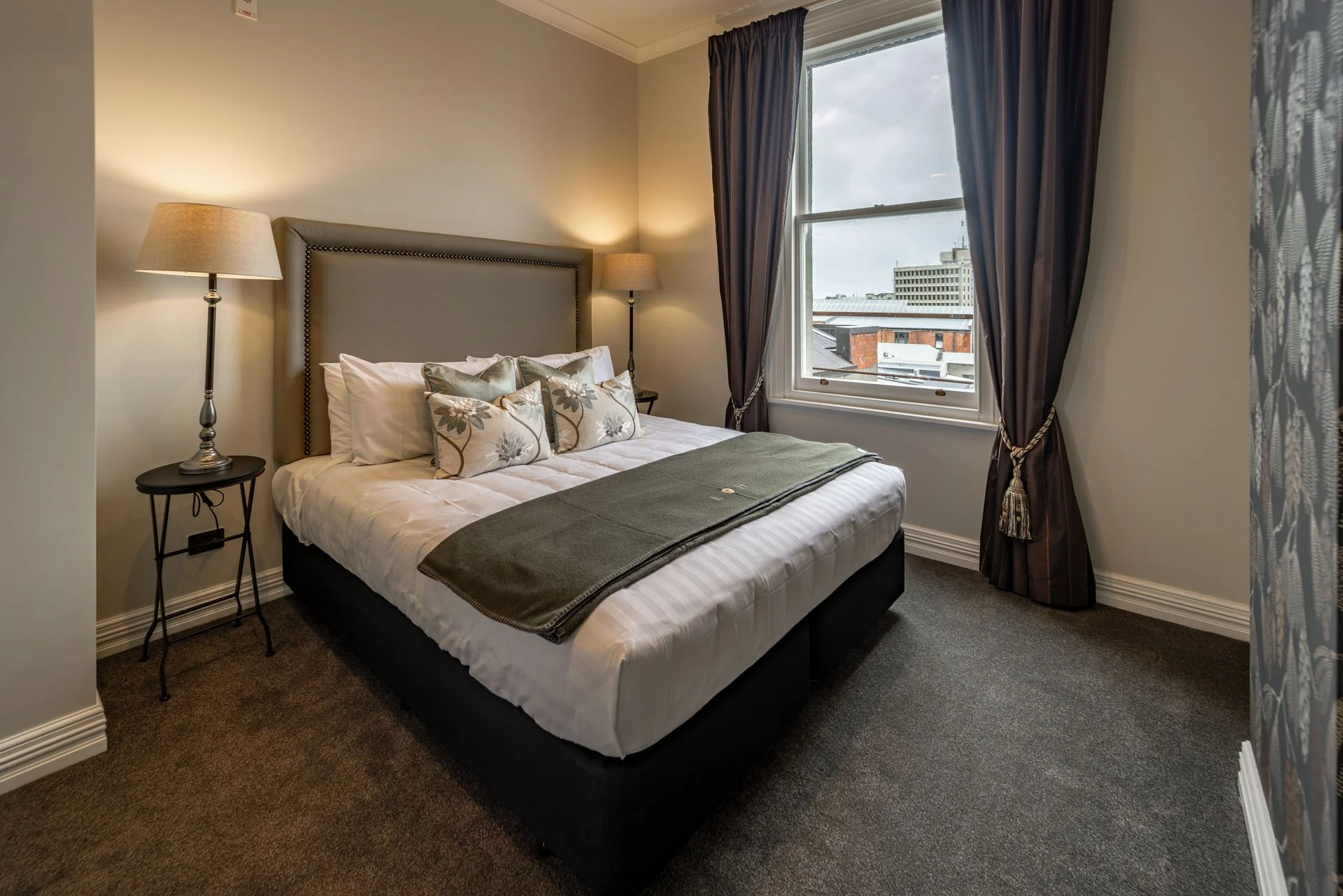 A bedroom with a bed, two table lamps, and a window with purple curtains showing city buildings outside.