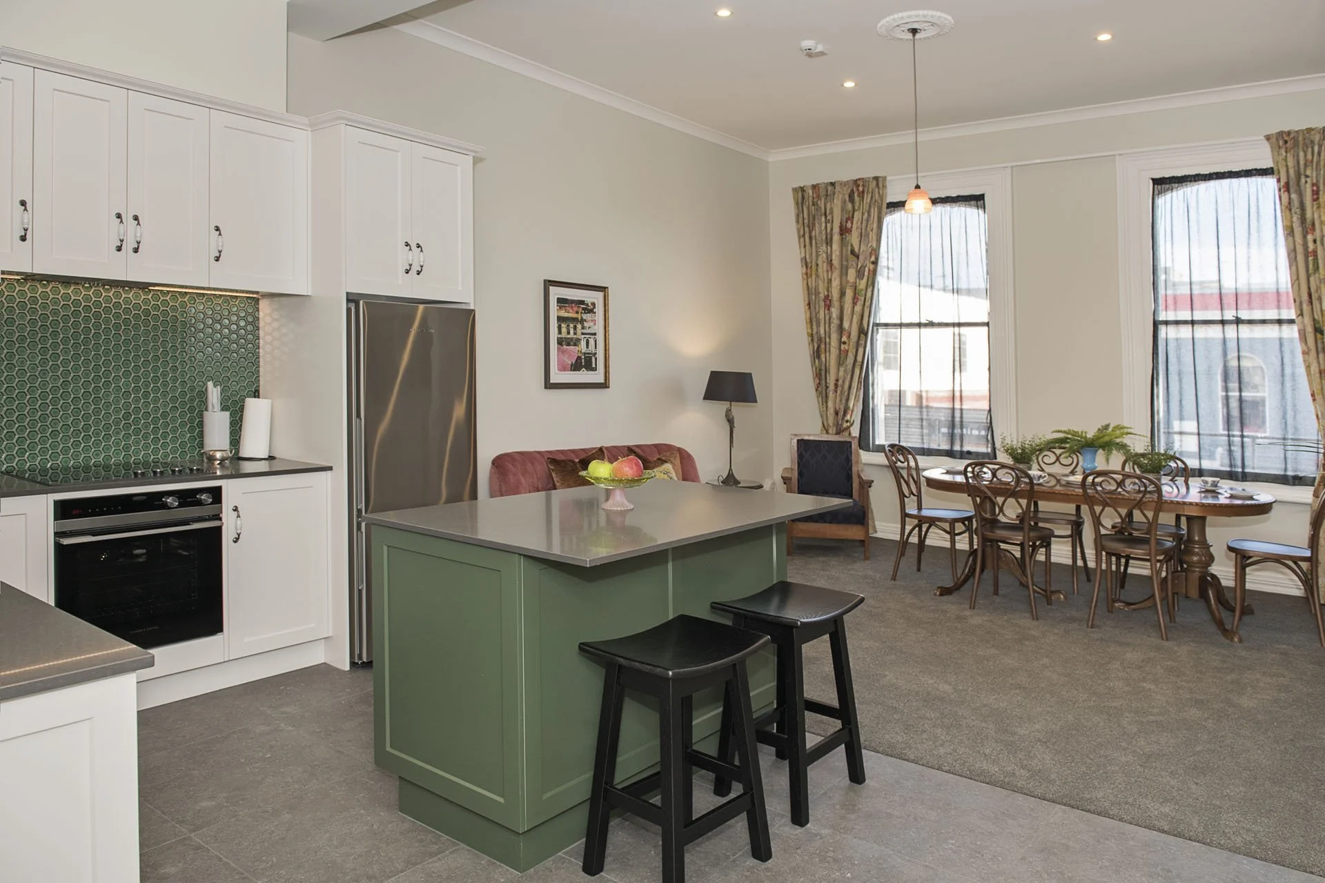 Open-concept kitchen and dining area with large windows, white cabinetry, and a green kitchen island with two black stools.