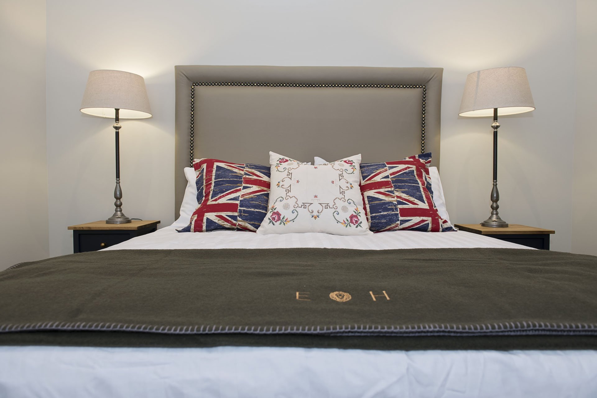 A neatly made bed with a gray upholstered headboard, decorated with Union Jack pillows and a white embroidered pillow, flanked by two nightstands with lamps, and a dark bedspread with initials embroidered on it.