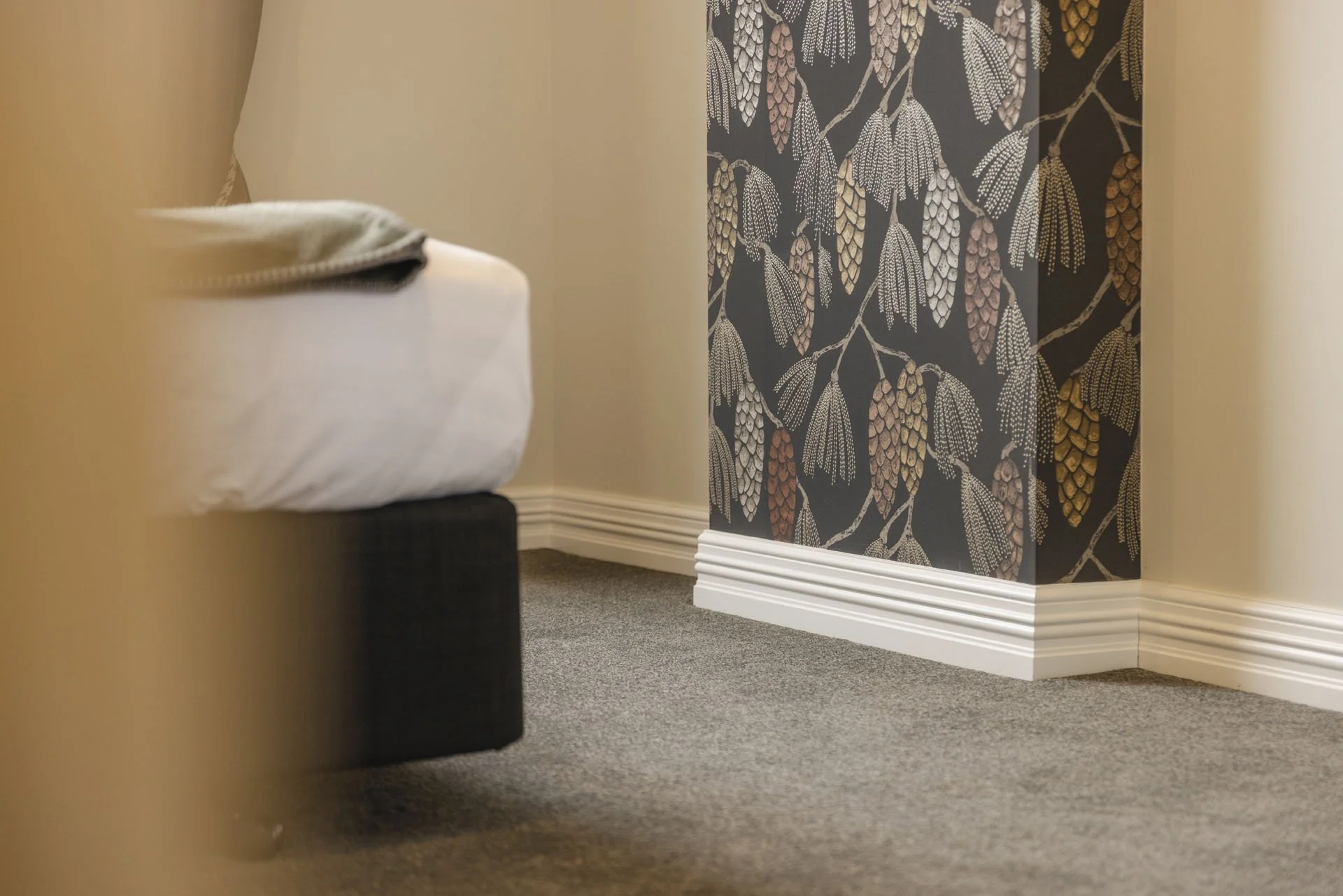 A corner of a room with beige walls, a dark patterned wallpaper with gold and white details on one wall, white baseboard, and beige carpet. Part of a bed with white bedding and a folded blanket is visible.