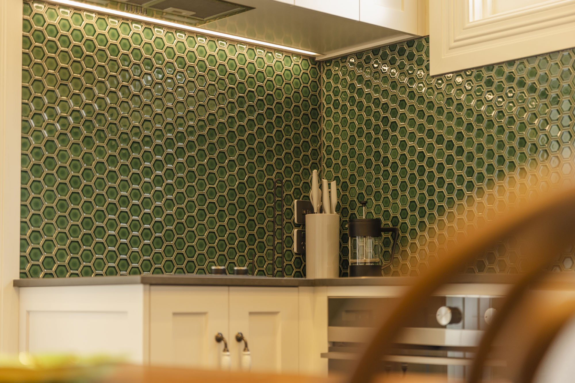 Kitchen with green honeycomb tile backsplash, white cabinets, a knife holder with knives, a coffee maker, and a portion of a chair in the foreground.