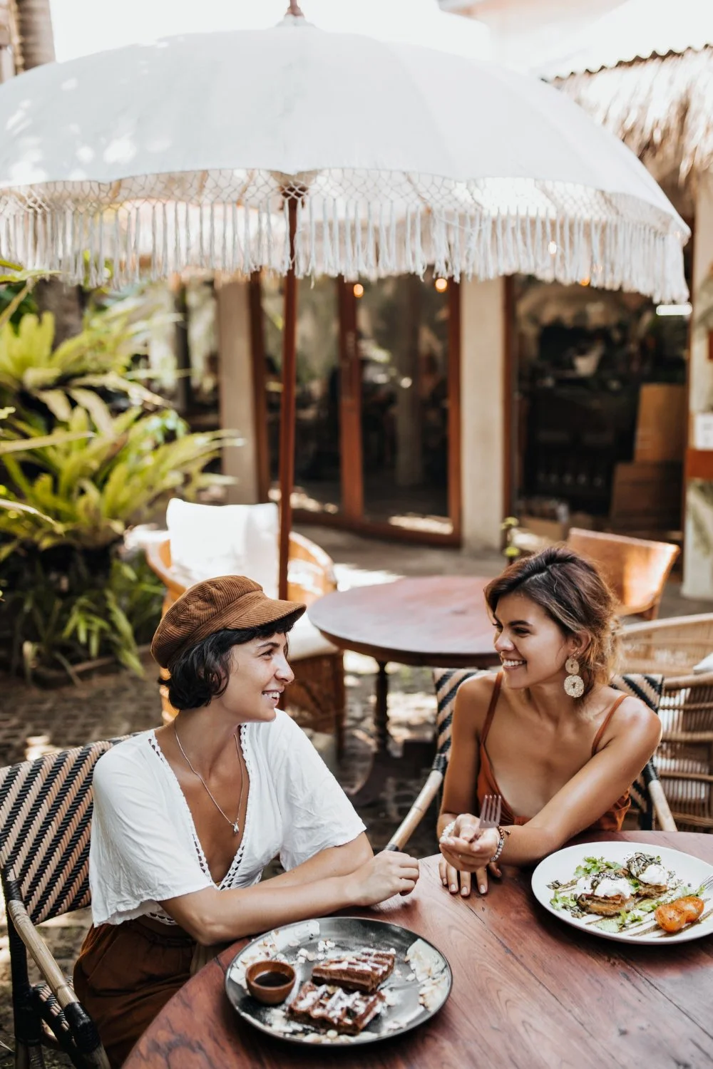 Two women sitting at an outdoor table under a white parasol, smiling and talking. They have plates of food in front of them, with one woman holding a fork. Lush plants surround the area, and there is a rustic building in the background.