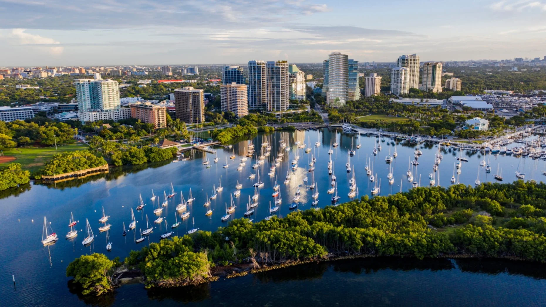 A cityscape with high-rise buildings, a marina filled with sailboats, and green trees along the water.