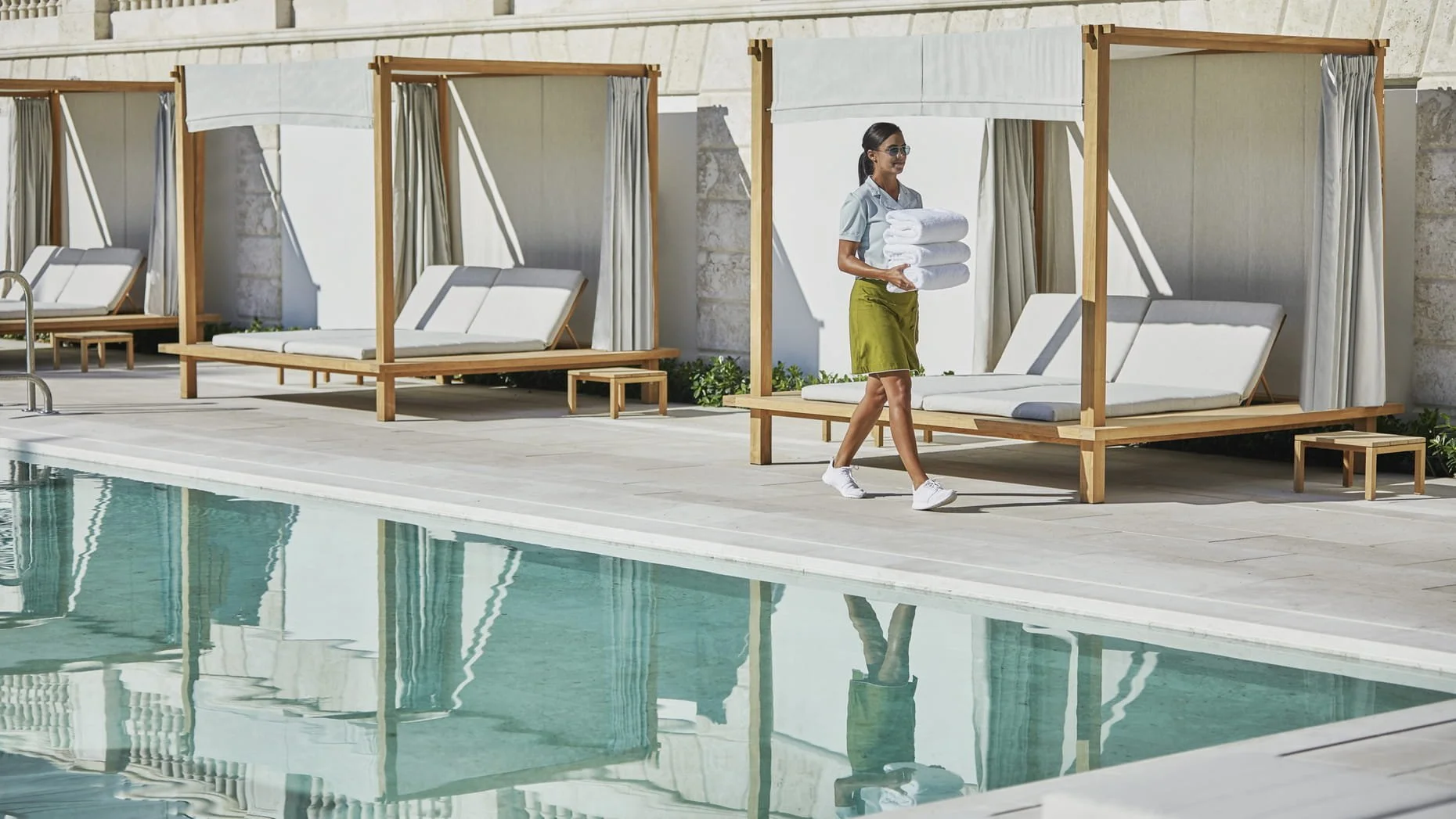 A woman in a light blue shirt and green shorts carrying white towels walking beside a pool with cabanas and lounge chairs.