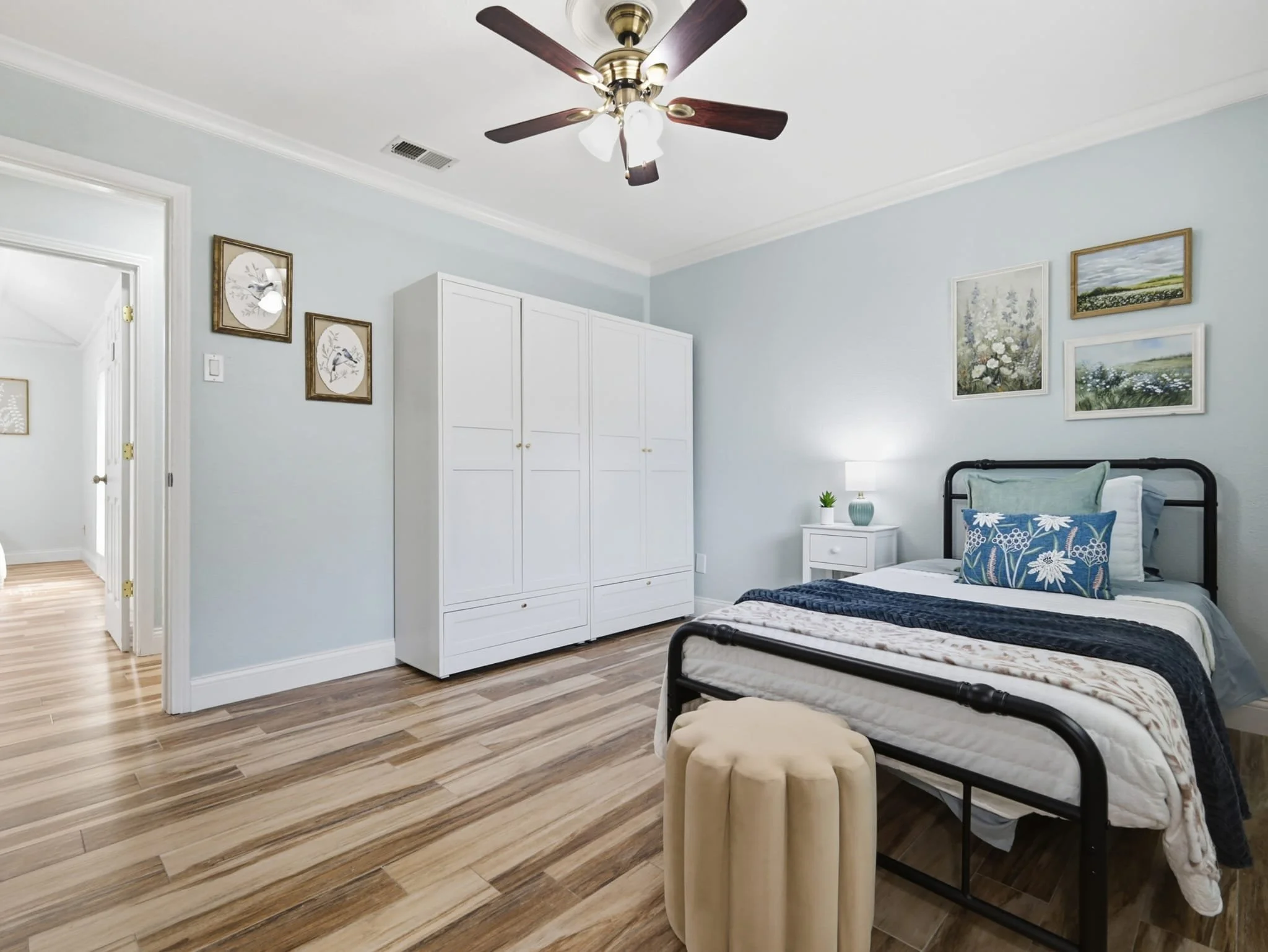 A bedroom with light blue walls, a black metal bed with blue and white bedding, a white bedside table with a lamp and a small plant, a white wardrobe, framed artwork on the walls, a ceiling fan, and a beige ottoman at the foot of the bed.