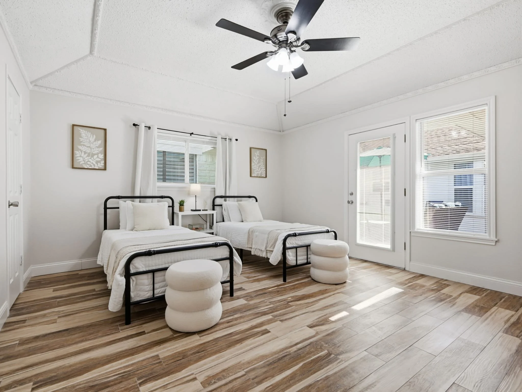 A bright bedroom with two twin beds with black metal frames, white bedding, and pillows. There are small white nightstands with lamps and a potted plant between the beds. The room has white walls with framed botanical art, a window with white blinds 