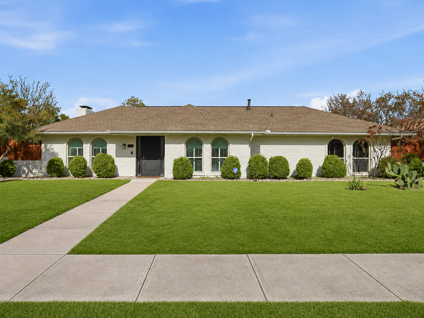 Front view of a single-story white house with a brown roof, a black front door, three arched and three rectangular windows with shutters, green bushes along the facade, a well-maintained lawn, and a concrete sidewalk leading to the front door under a