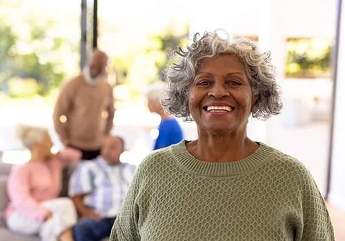 Smiling elderly woman with curly gray hair in a green sweater, standing in a bright room with blurred people in the background.