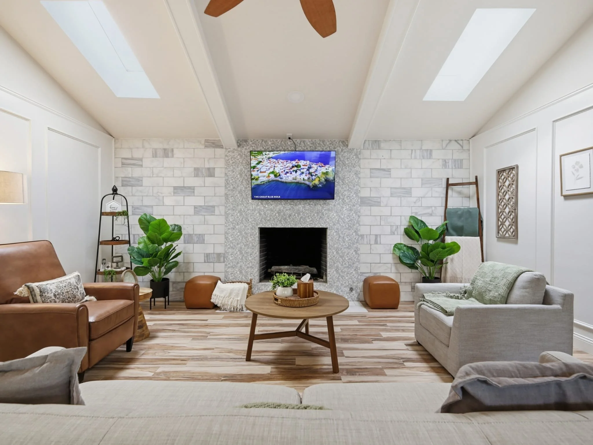 Living room with white brick fireplace, wall-mounted TV, skylights, ceiling fan, beige and gray sofas, wooden coffee table, green plants, framed wall art, and decorative ladder.