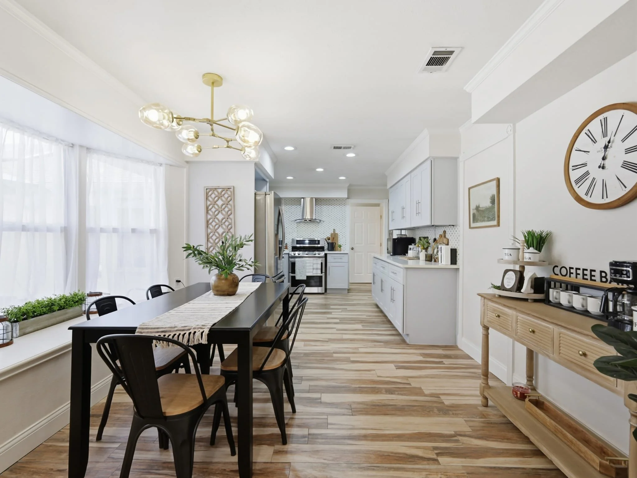 Bright kitchen and dining area with white cabinetry, wooden flooring, a black dining table with chairs, and a wall clock.