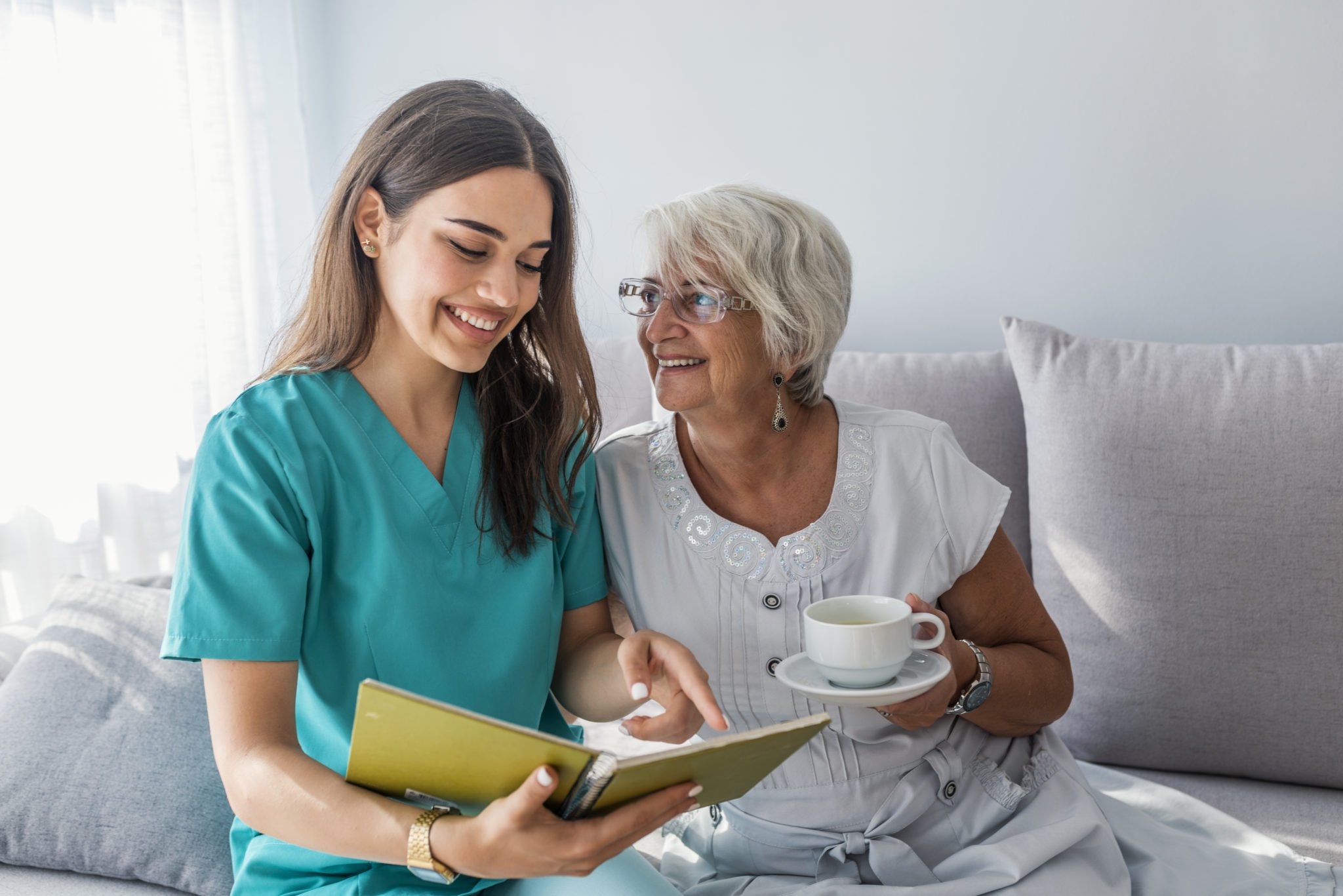 A young woman in medical scrubs and an elderly woman sitting on a couch, smiling and sharing a moment. The elderly woman is holding a cup of tea or coffee, and the young woman is looking at a notebook or journal.
