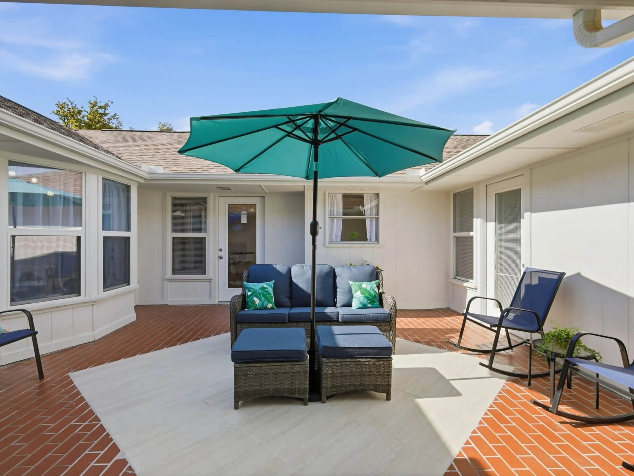 Outdoor patio with blue seating, large teal umbrella, and rocking chairs under a clear blue sky.