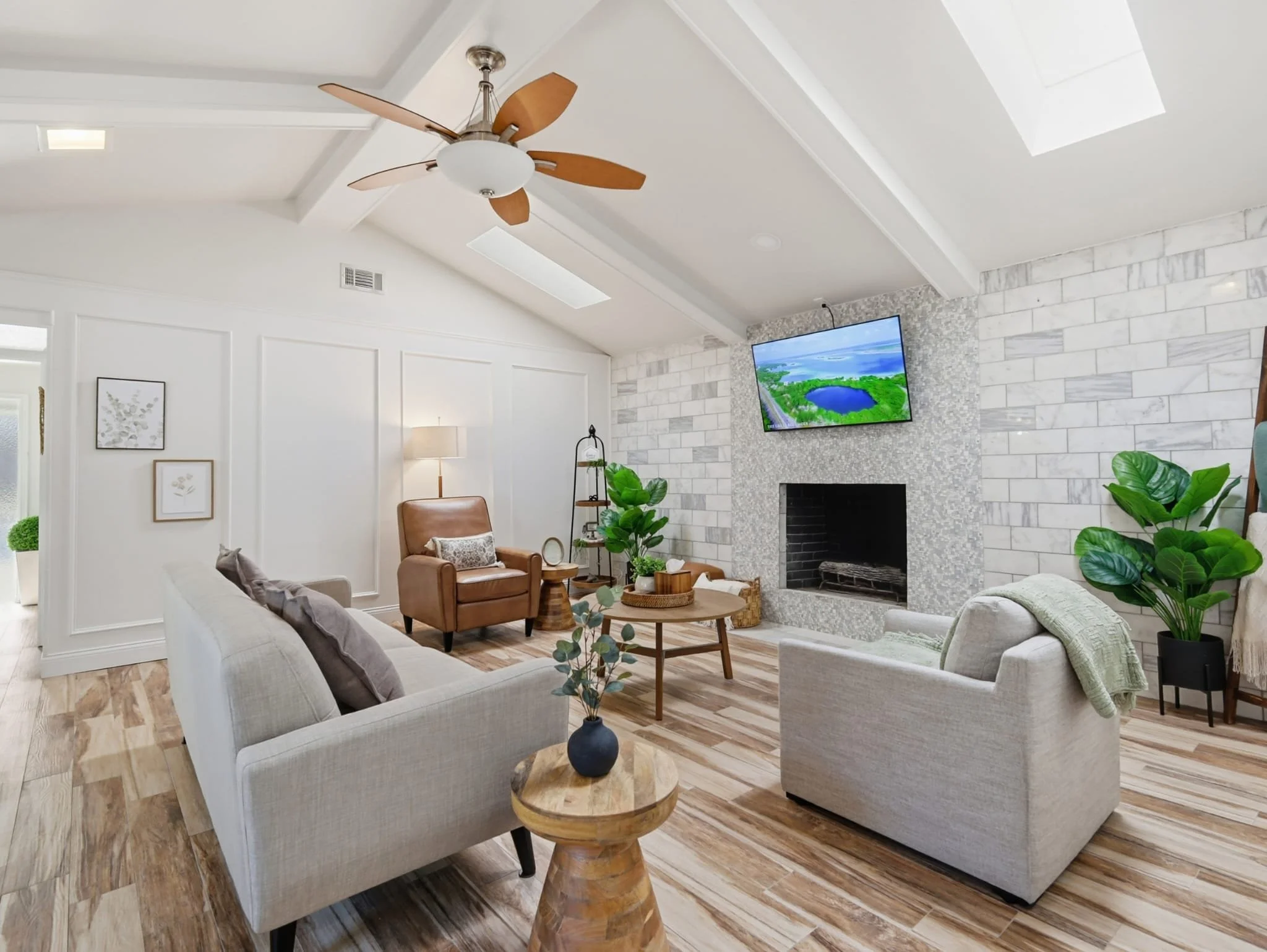 Modern living room with white walls, a fireplace, a flat-screen TV, and multiple sofas and armchairs. Decor includes potted plants, framed artwork, and a wooden coffee table. Recessed lighting and a skylight illuminate the space.
