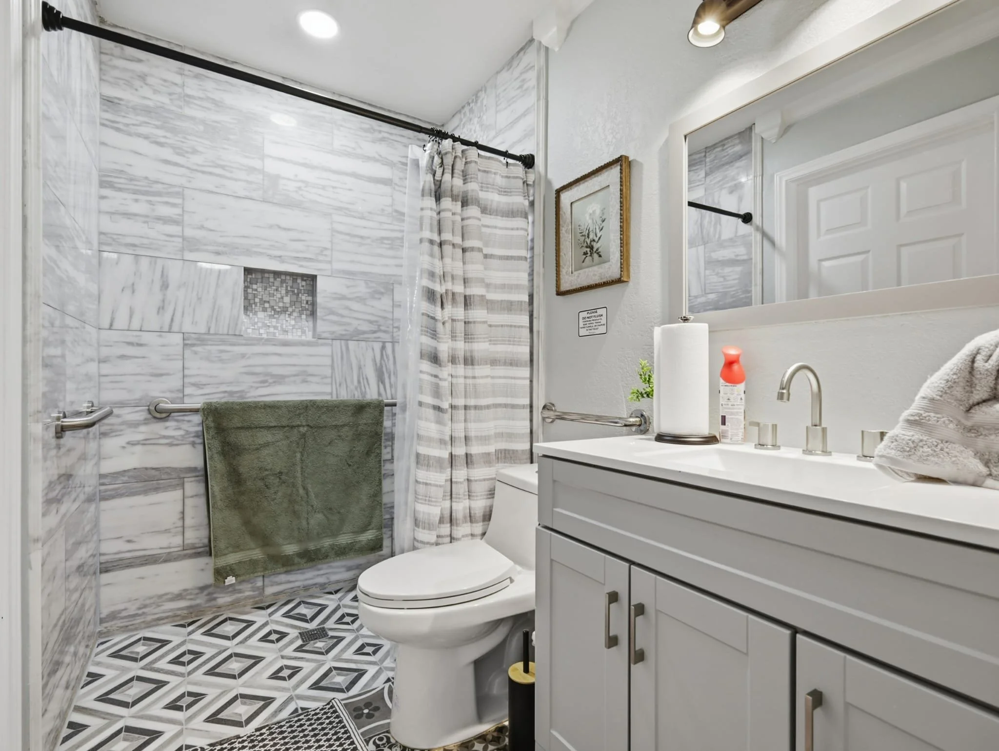 A modern bathroom with a walk-in shower featuring marble tiles, a green towel on a horizontal bar, and a striped shower curtain. The vanity has a white countertop with a sink, a soap dispenser, a paper towel roll, and a small green plant. A large mir