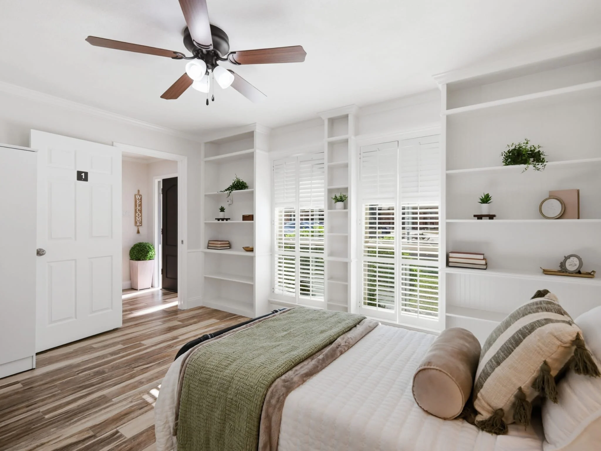Bright bedroom with white walls, wooden flooring, a bed with beige and green bedding, and decorative pillows. Large windows with white shutters, white built-in shelves with plants, books, and decorative items. Ceiling fan with wooden blades and light