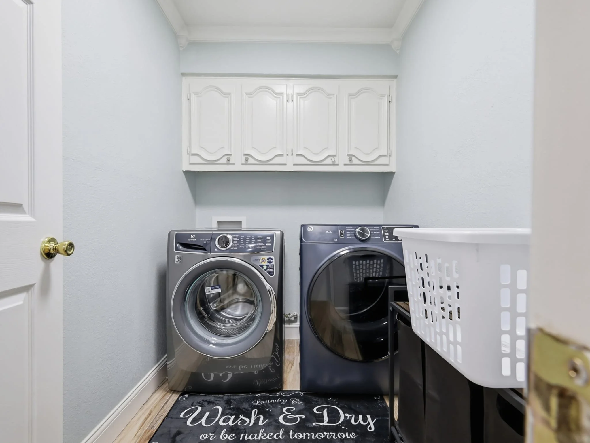A laundry room with a front-loading washing machine and dryer, white cabinets above, a laundry mat on the floor reading "Wash & Dry or be naked tomorrow," and laundry supplies on a black table.
