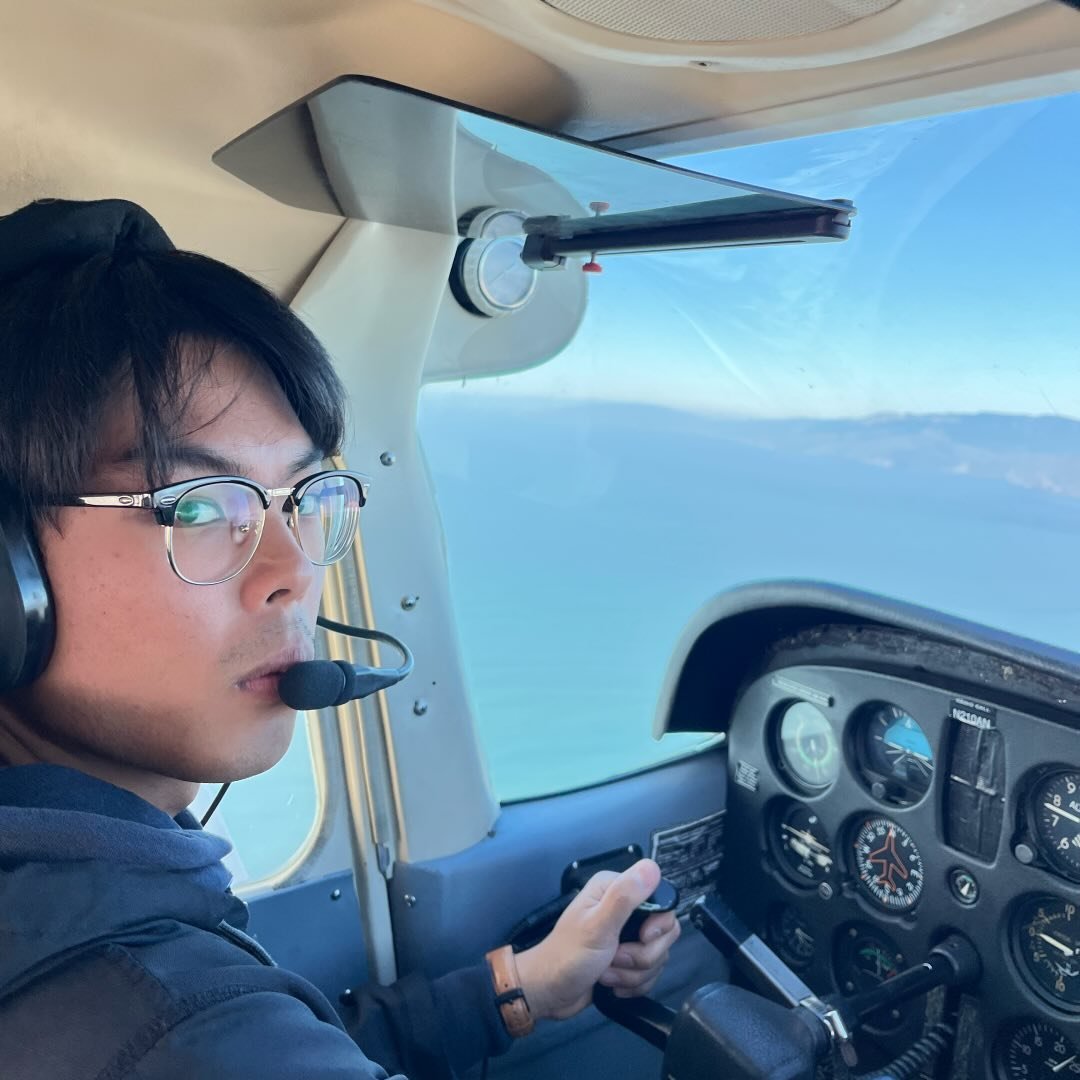 A young man flying a small aircraft, wearing glasses and a headset, with a view of the ocean and sky visible through the cockpit window.