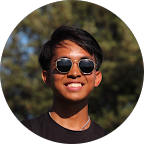 A smiling person with short dark hair and sunglasses outdoors on a sunny day.