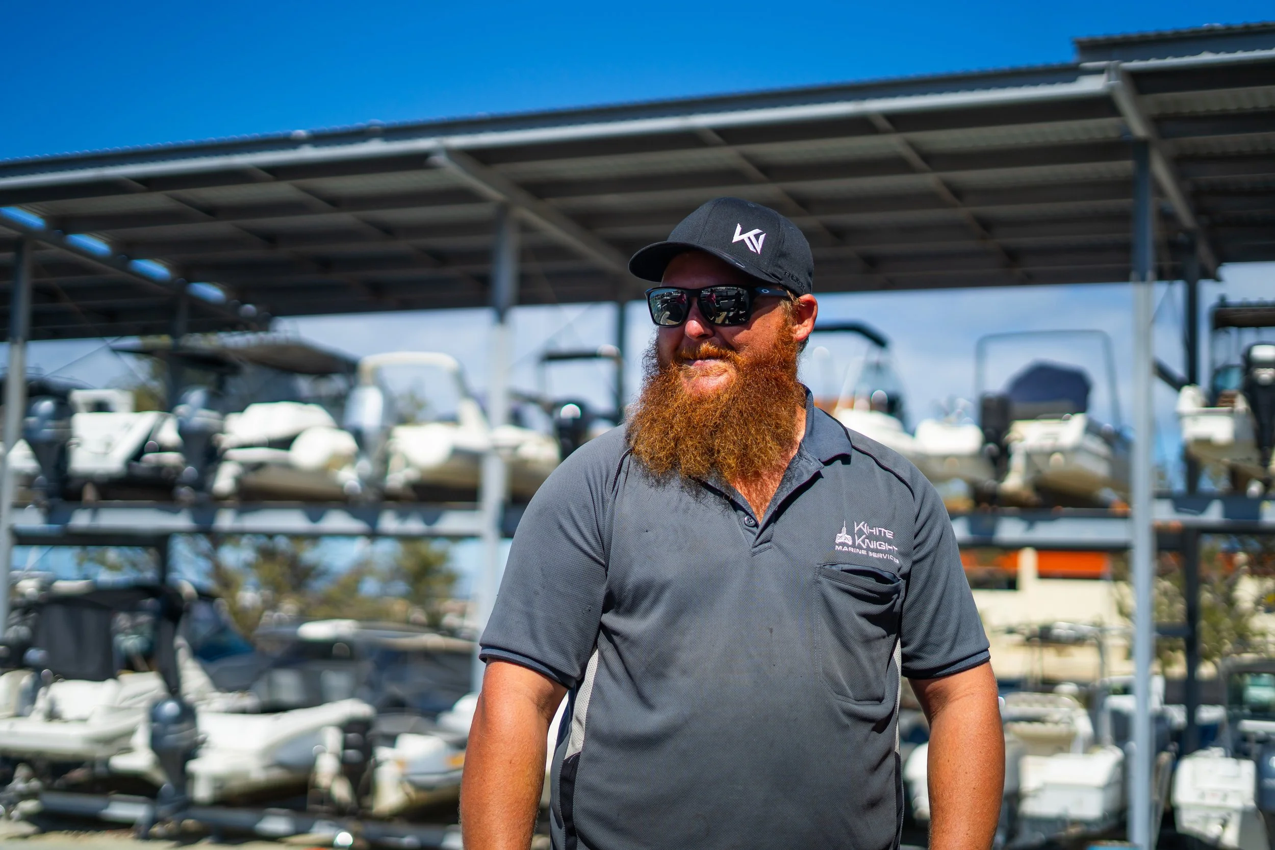 A man with a red beard wearing sunglasses and a black cap, standing outdoors near a marina with boats and a blue sky in the background.