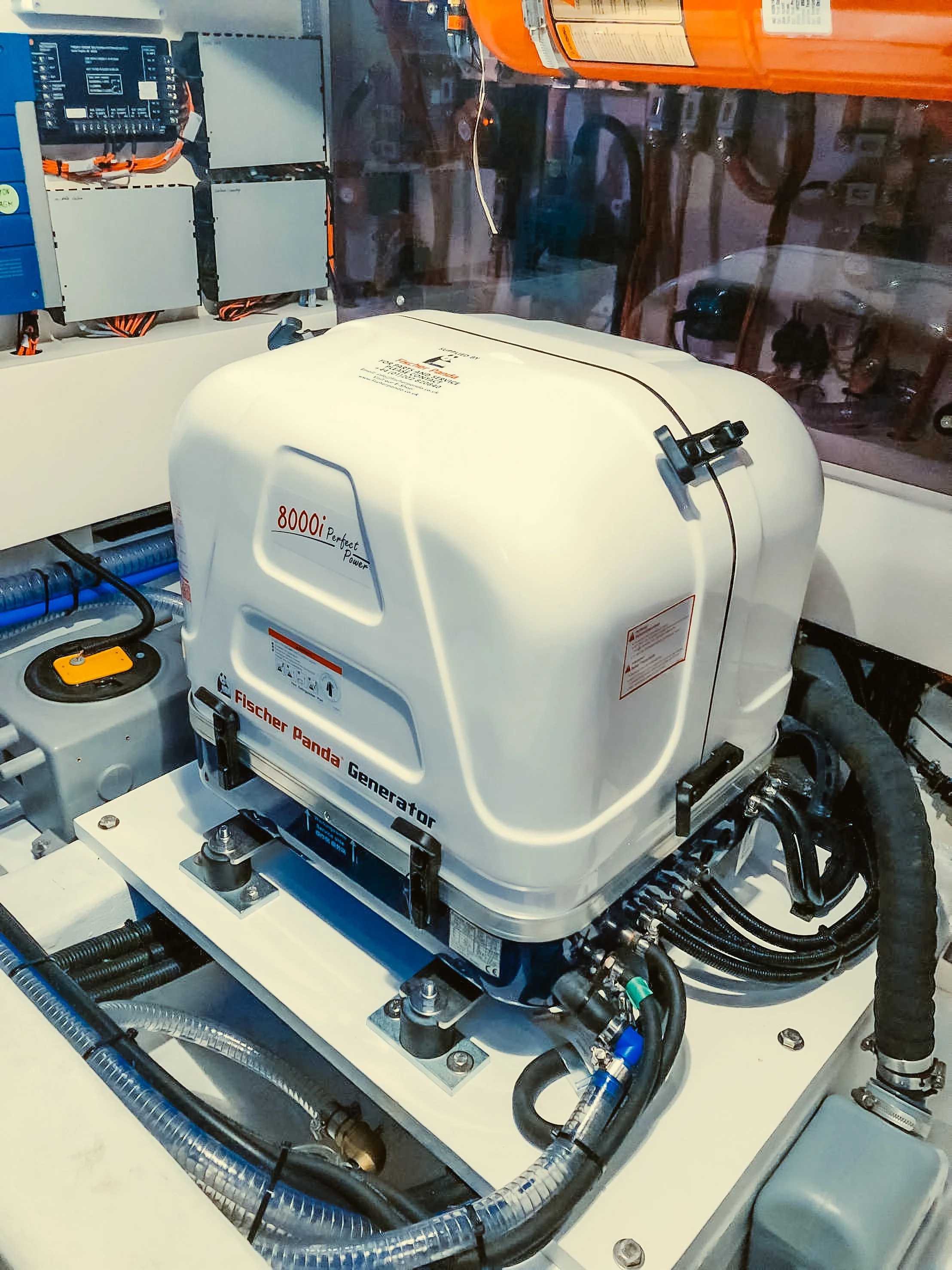 Close-up view of a generator with a white casing, surrounded by various electrical components, wires, and tubing, inside a mechanical or industrial setting.
