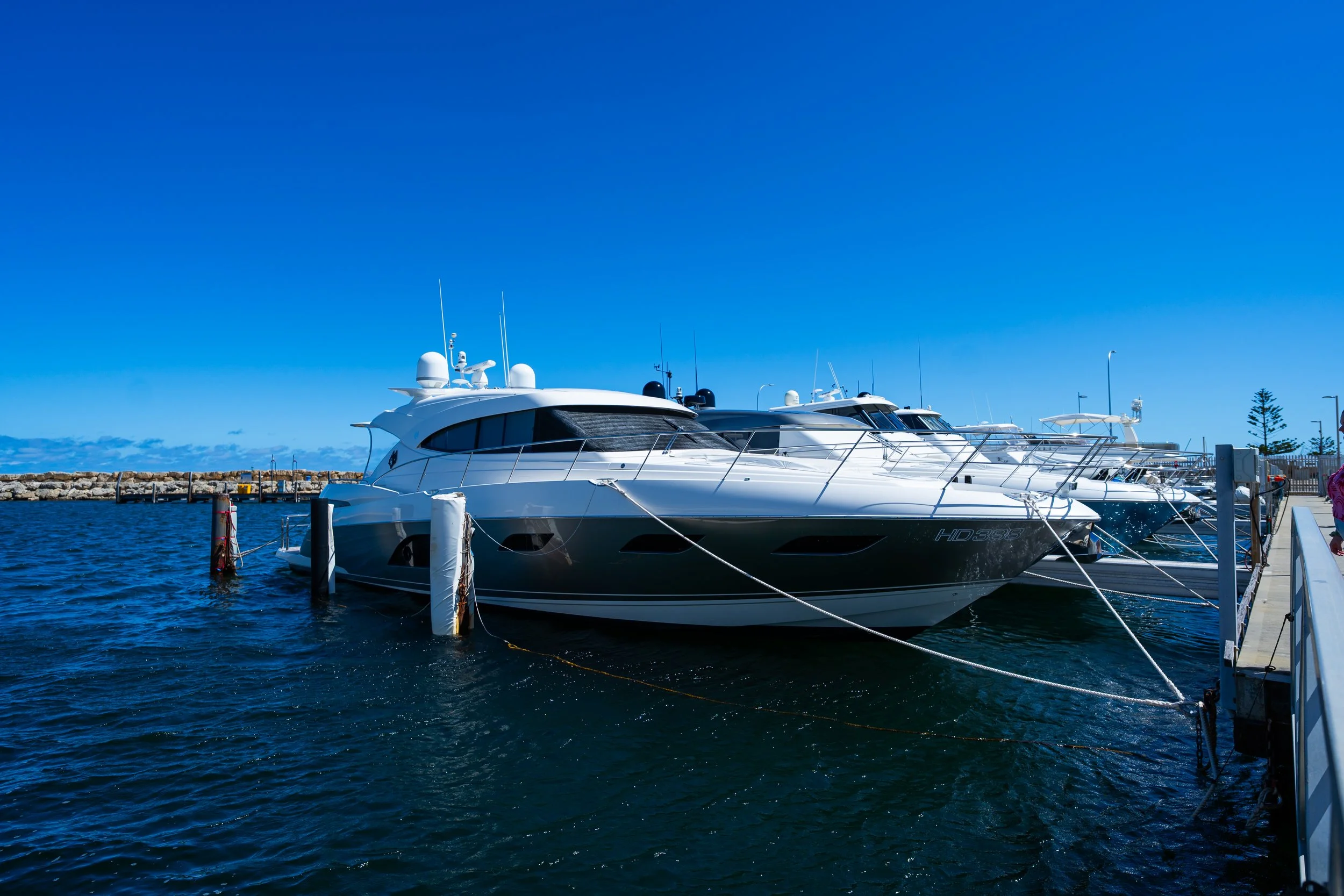Several luxury yachts docked at a pier on a clear, sunny day with a blue sky and calm water.
