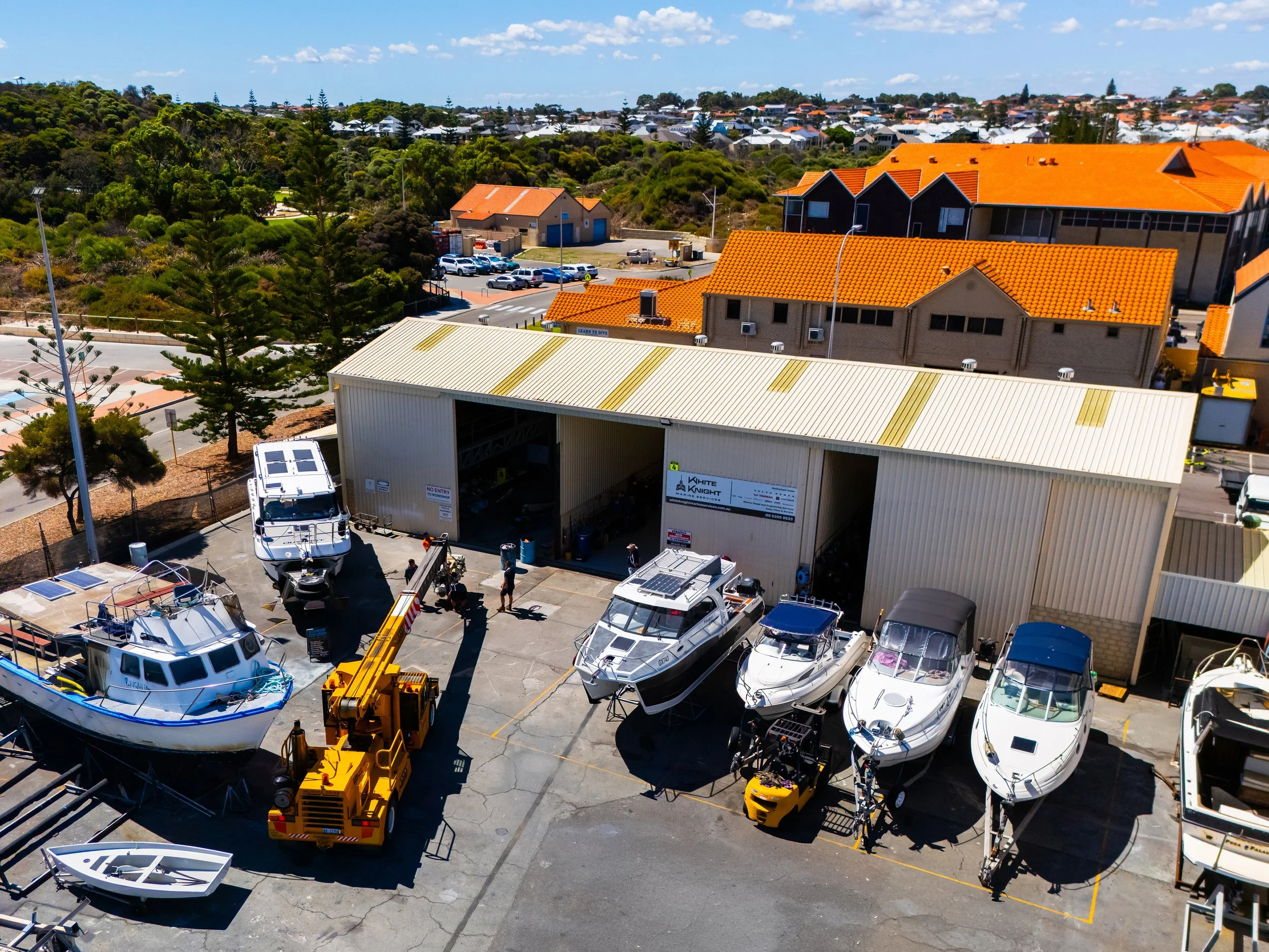 An outdoor marina or boatyard with several boats docked and equipment for boat maintenance. In the background, a residential area with houses and greenery on hills under a clear blue sky.