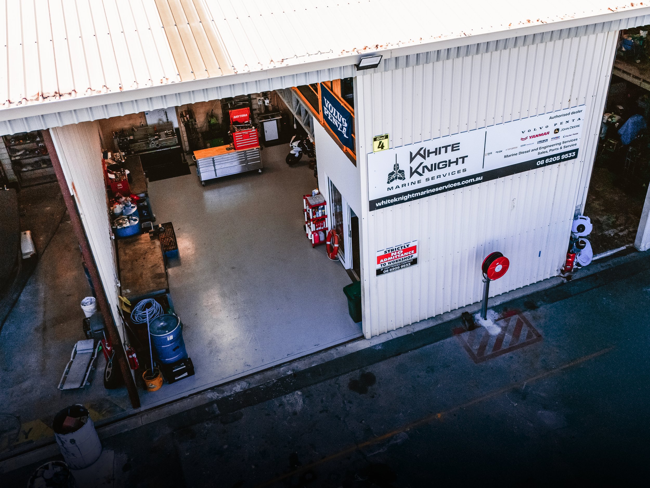Aerial view of a garage or workshop with white corrugated metal walls, open large sliding door, containing tools, a motorcycle, and equipment. Signage reads 'White Knight Marine Services' and other business information.