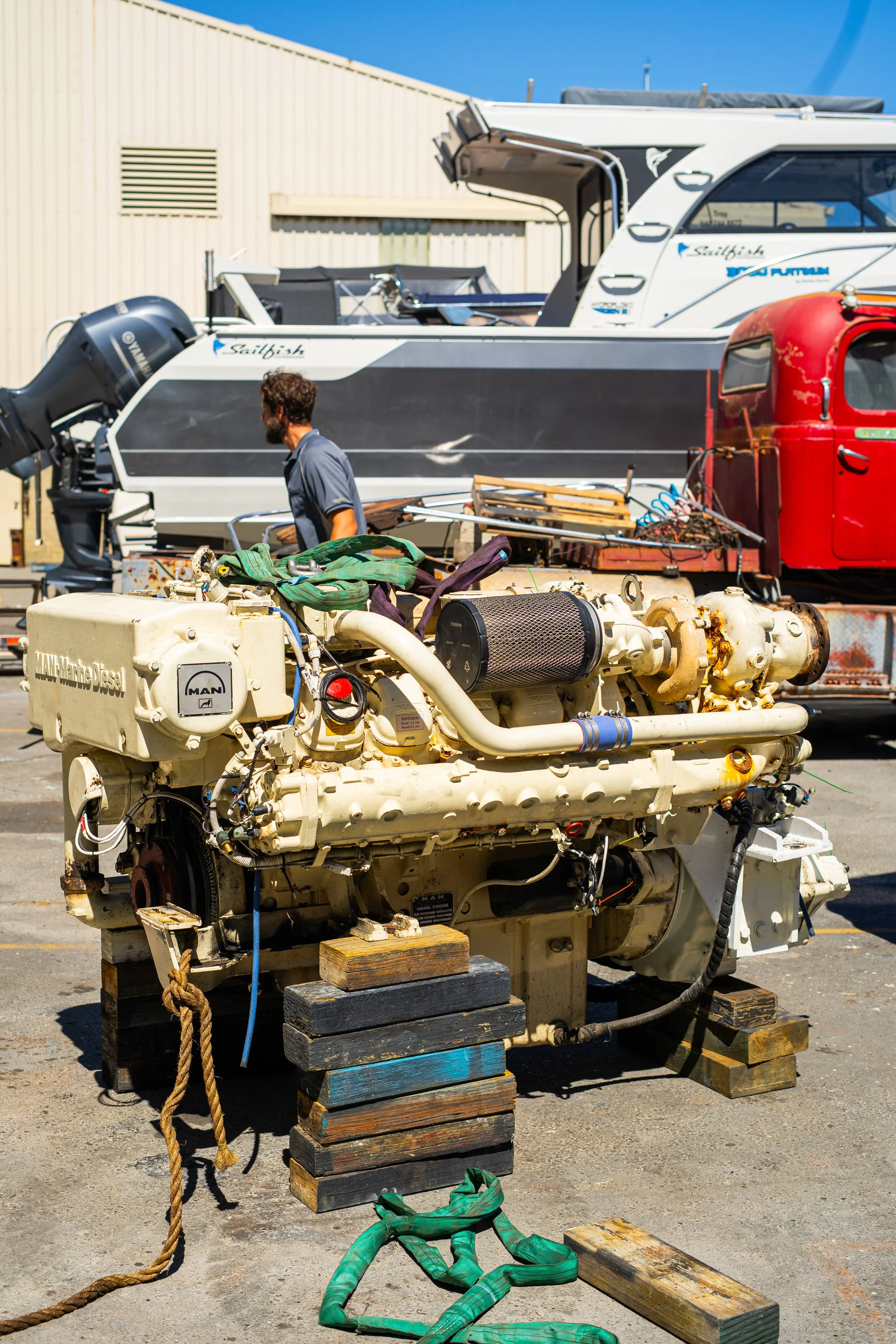 A large marine diesel engine on wooden supports, with a man working nearby, and boats in the background.