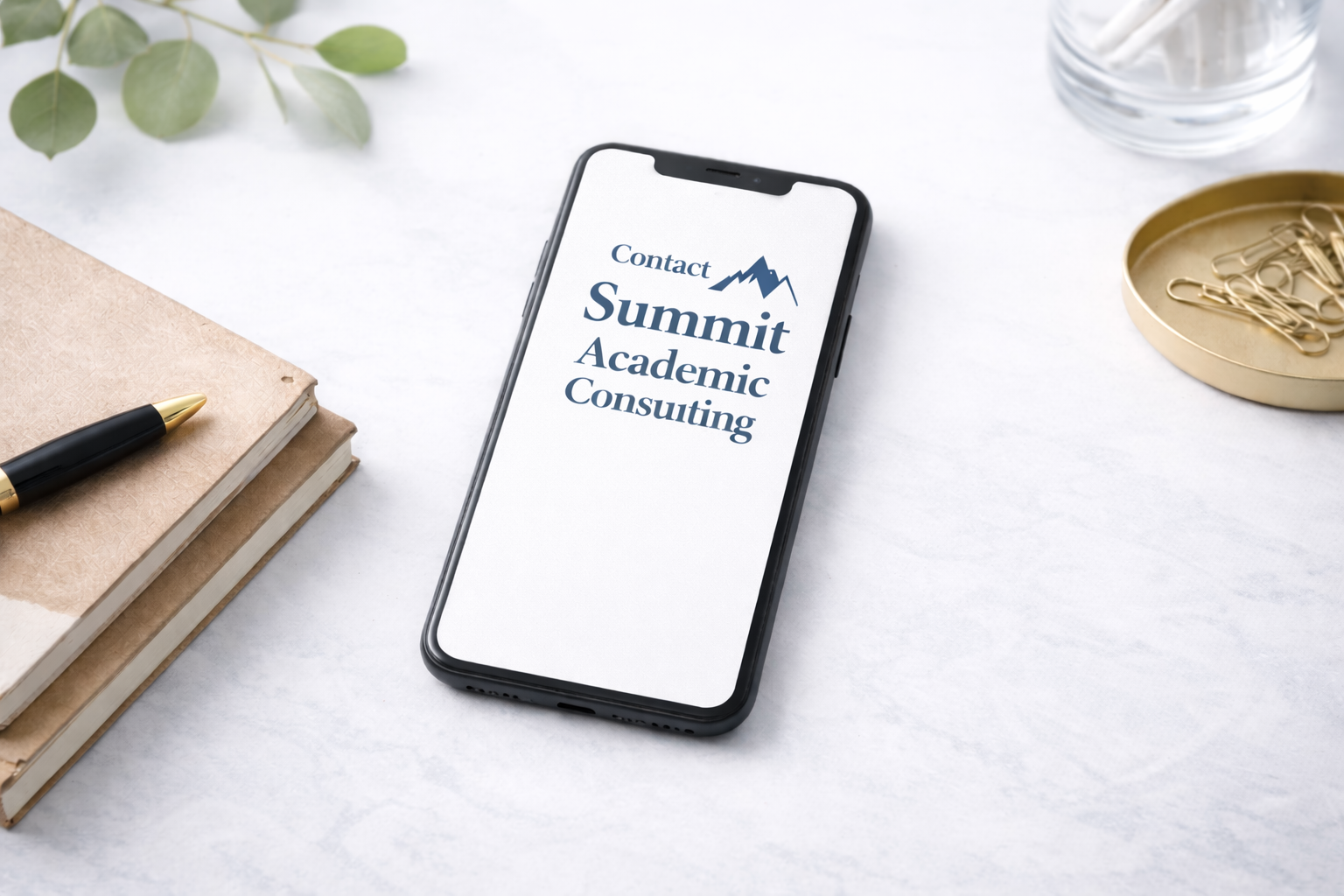 Smartphone on white desk displaying Summit Academic Consulting logo, nearby beige notebooks, pen, gold paper clips, glass of water, green leaves.