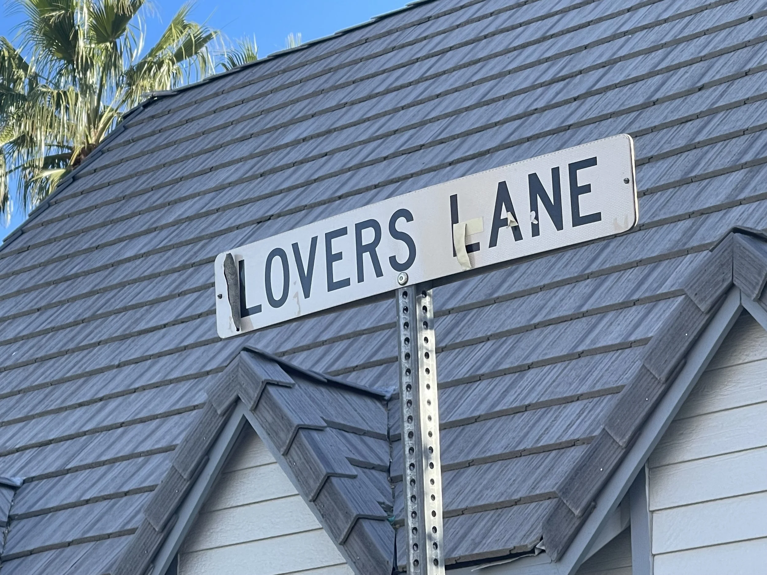 Street sign that reads 'Lovers Lane' mounted on a pole at a corner of a house with gray shingles and a blue sky in the background.