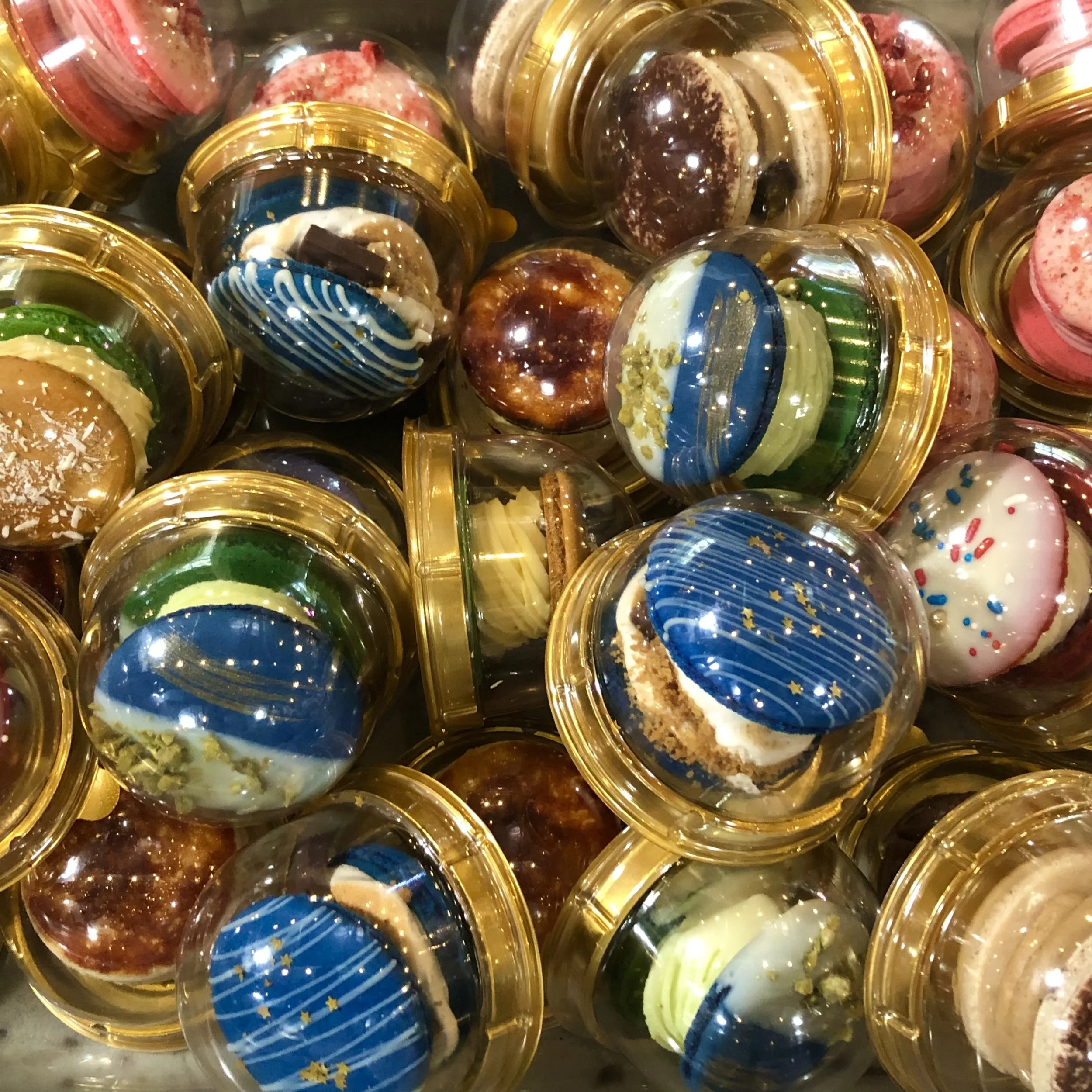 Assorted decorated cupcakes in small clear plastic containers with golden lids, featuring various colorful icing and sprinkle designs.