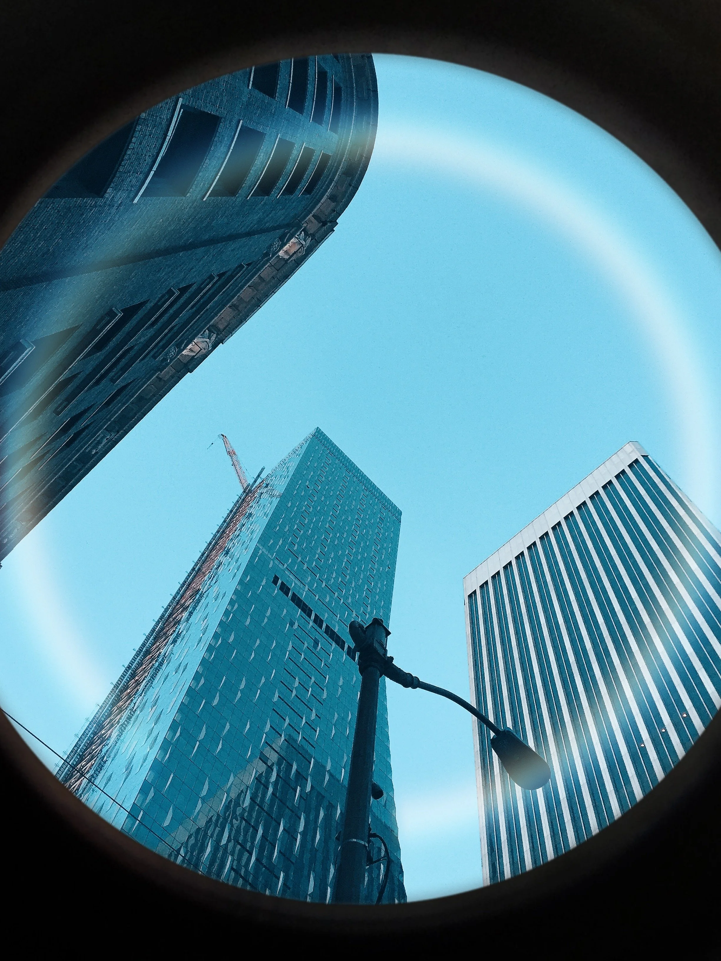 View of three tall modern skyscrapers with glass facades seen from below through a circular frame, with a streetlight in the foreground against a clear blue sky.