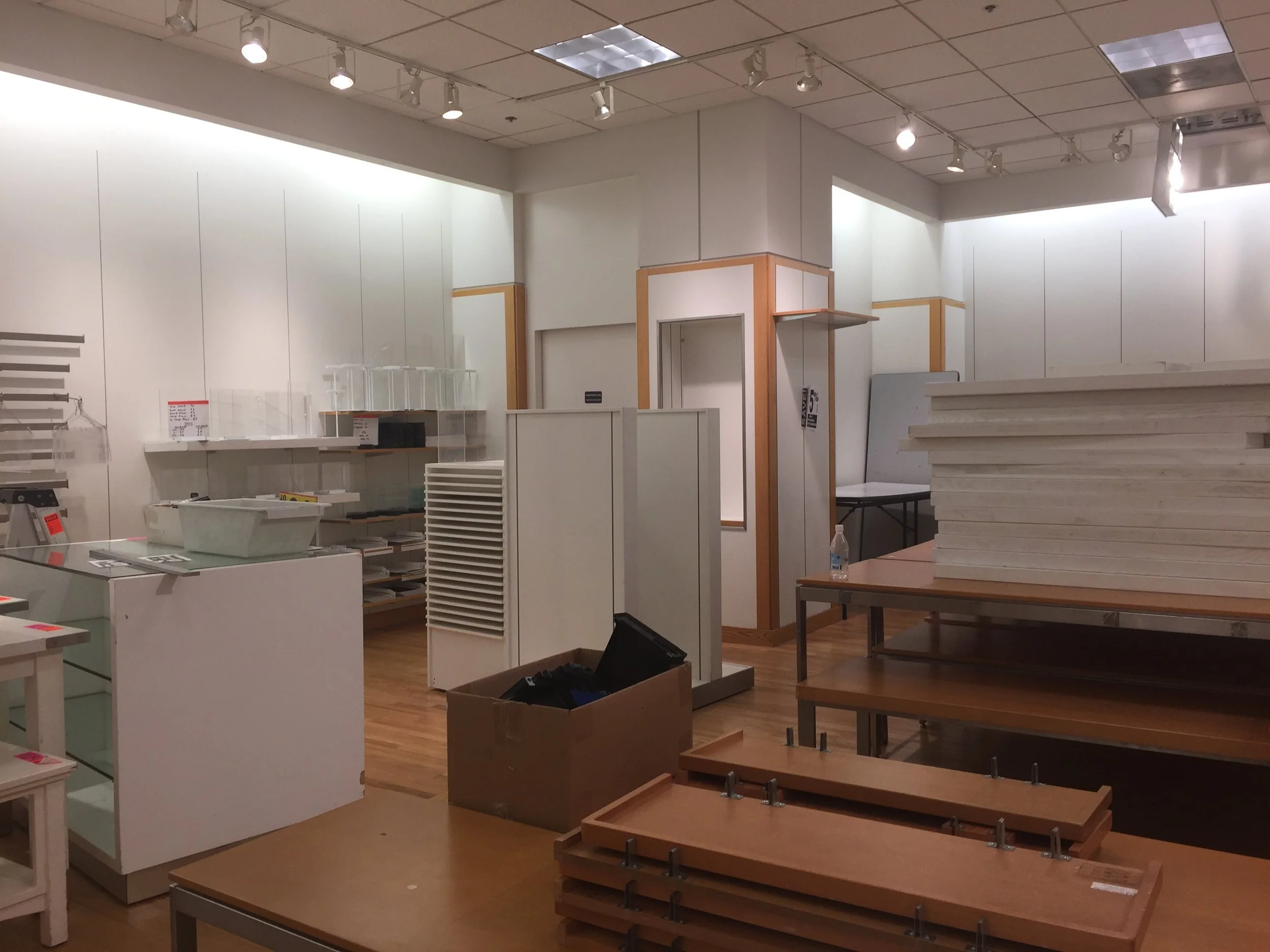 Empty retail store or display area with shelves, tables, and stacked white panels, with minimal items and a water bottle on one table.