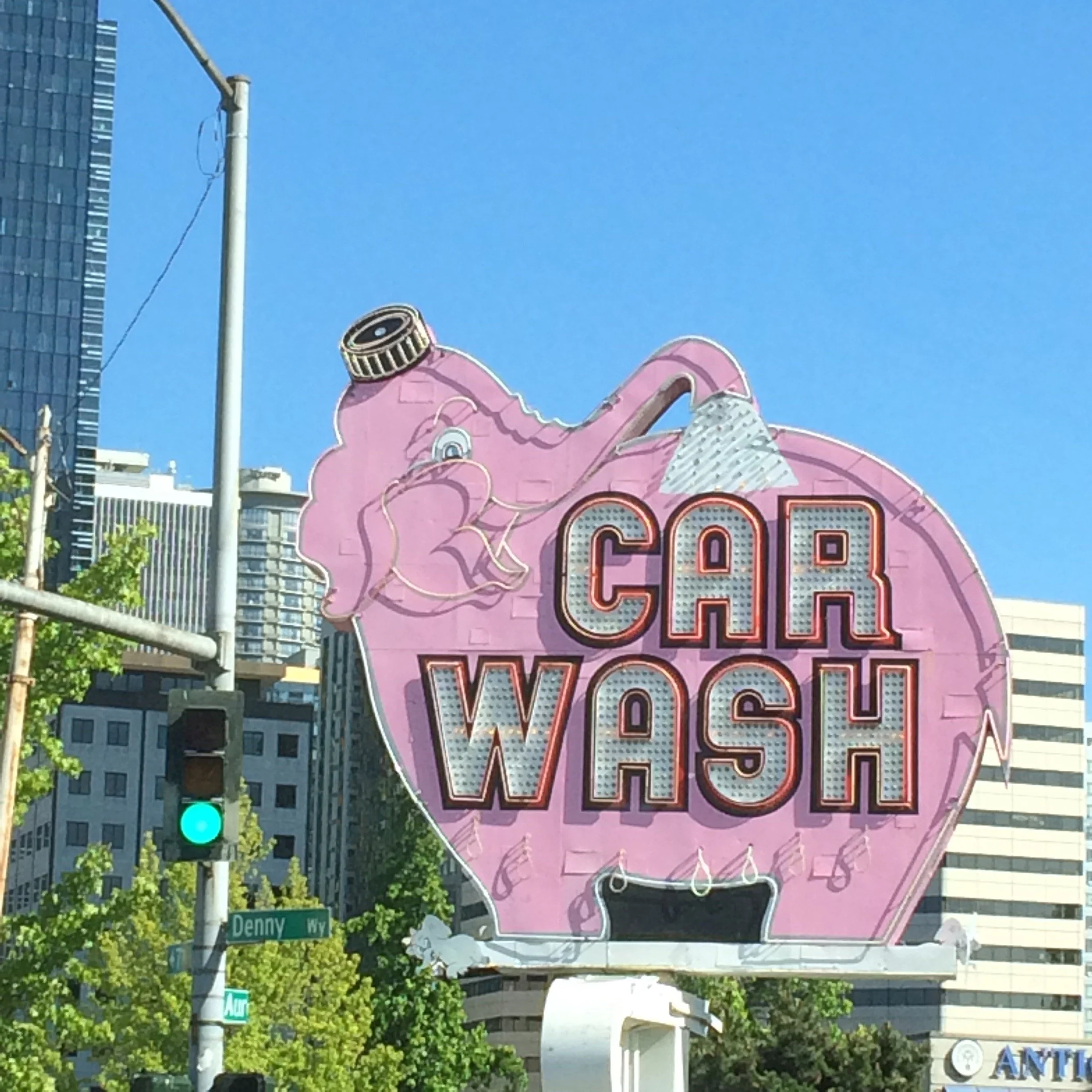 Neon sign shaped like a pink washing machine with the words "Car Wash" illuminated in red and white lights, located on a street corner against a backdrop of office buildings and trees under a clear blue sky.