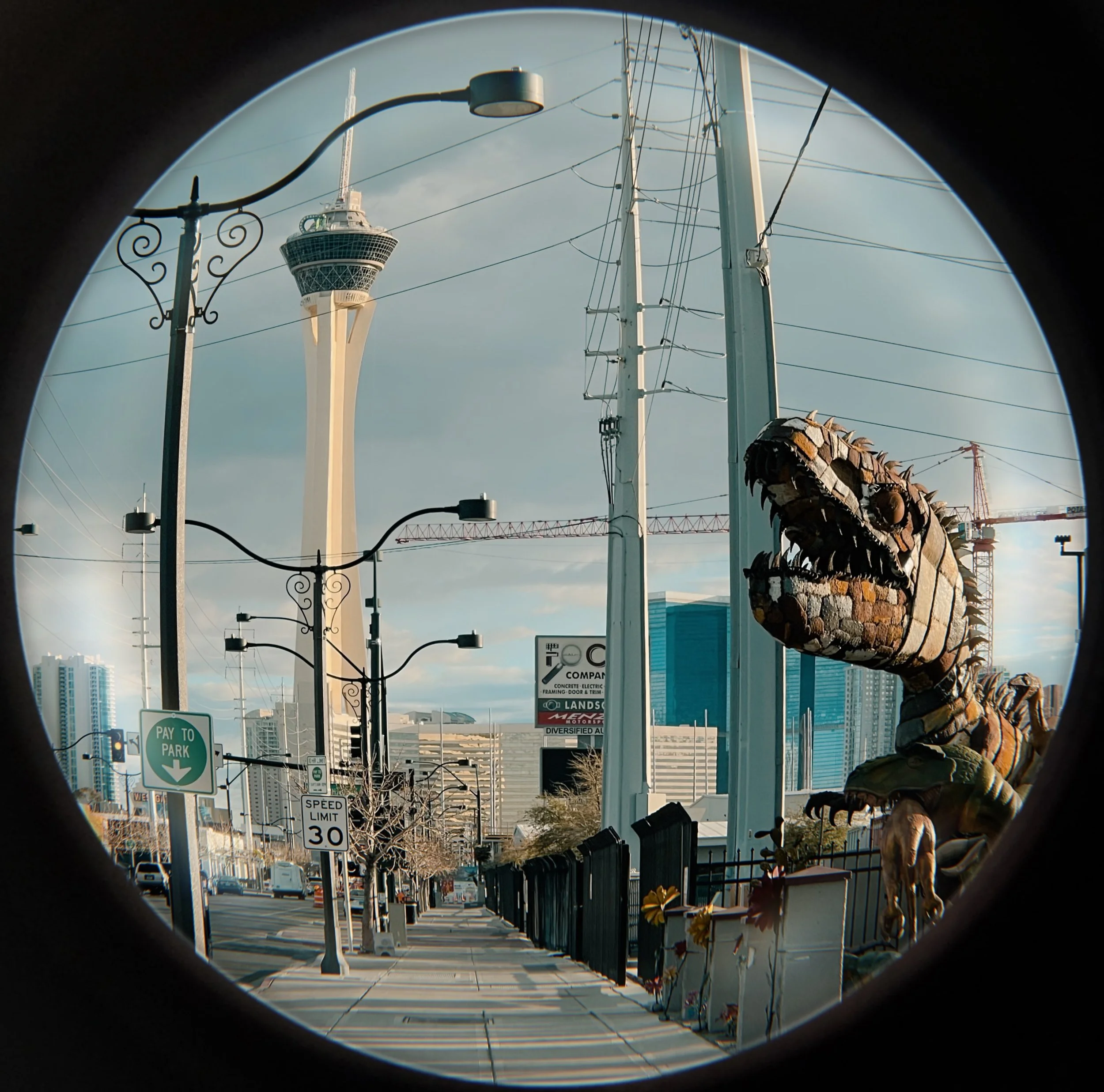 City street view with the Stratosphere Tower and downtown buildings in Las Vegas, including a large dinosaur sculpture on the sidewalk, seen through a circular frame.
