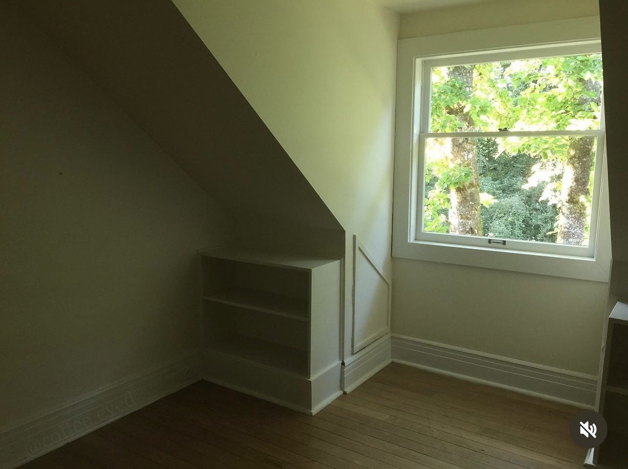 Empty room with sloped ceiling, a window with a view of green trees, and built-in white shelves.