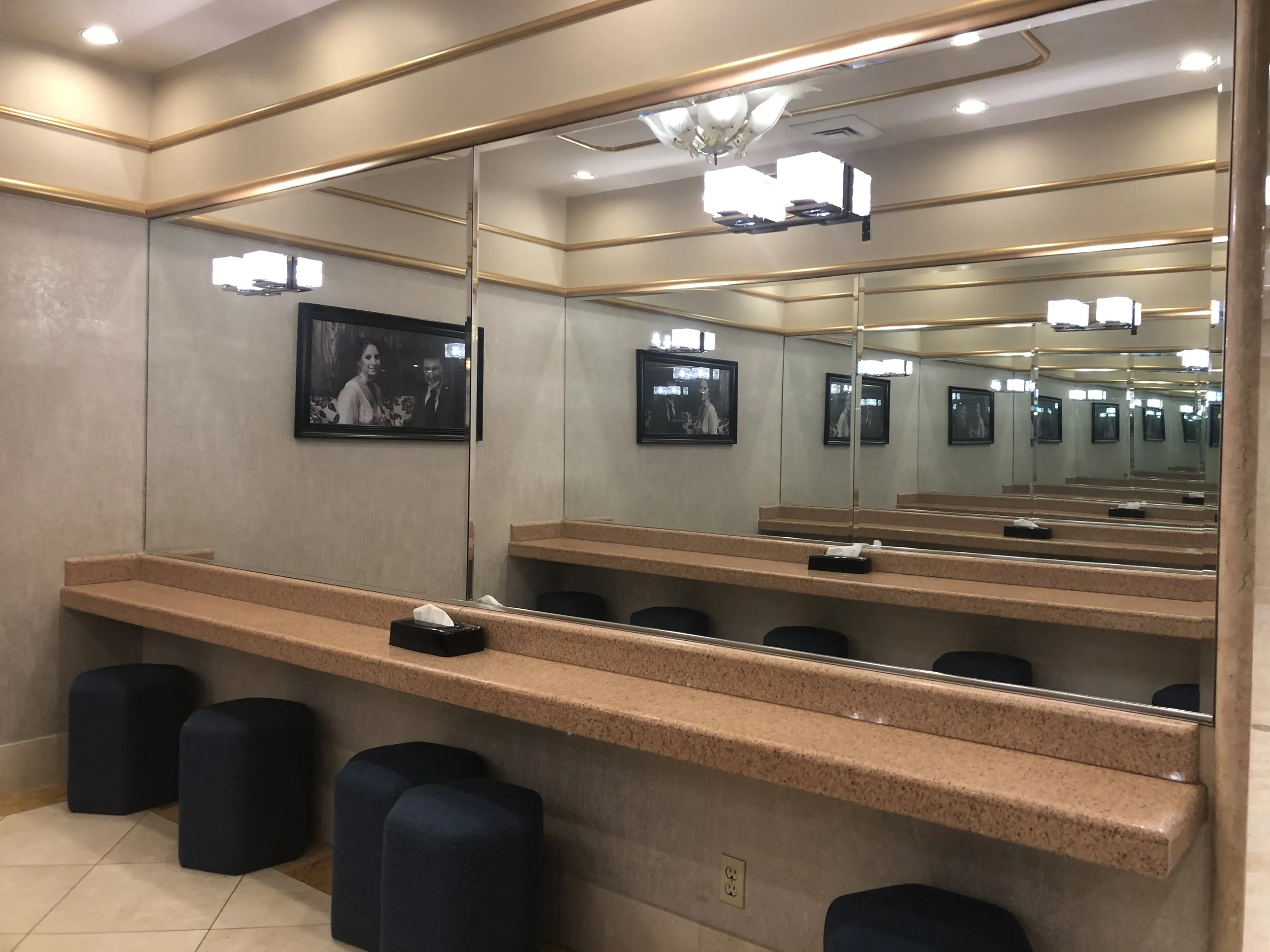 A dressing room with large mirrors on the wall, showing multiple reflections. There is a beige countertop with tissue boxes, black stools underneath, and framed black-and-white photographs on the wall. Ceiling lights illuminate the space.
