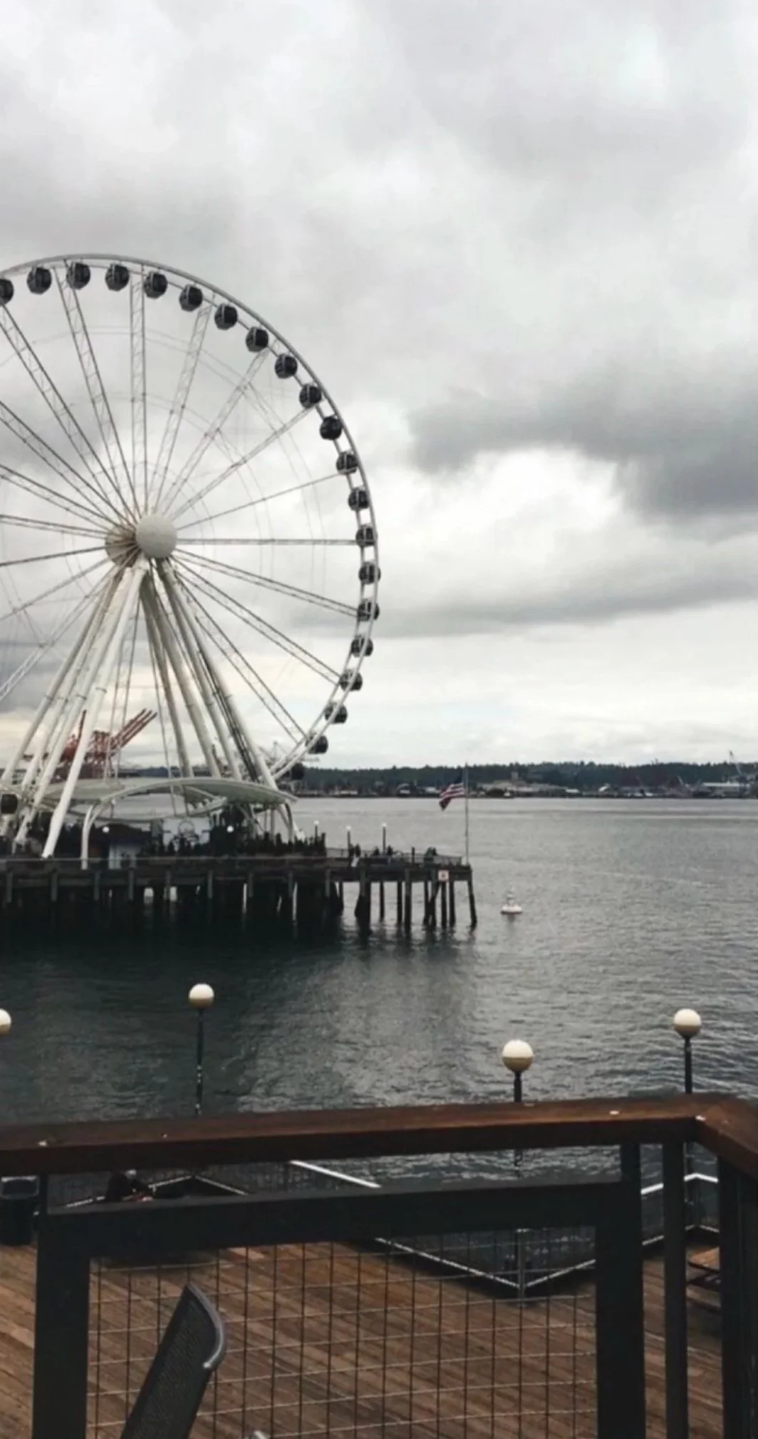 View of a waterfront with a large Ferris wheel on a cloudy day, boats on the water, and a wooden deck with outdoor seating.