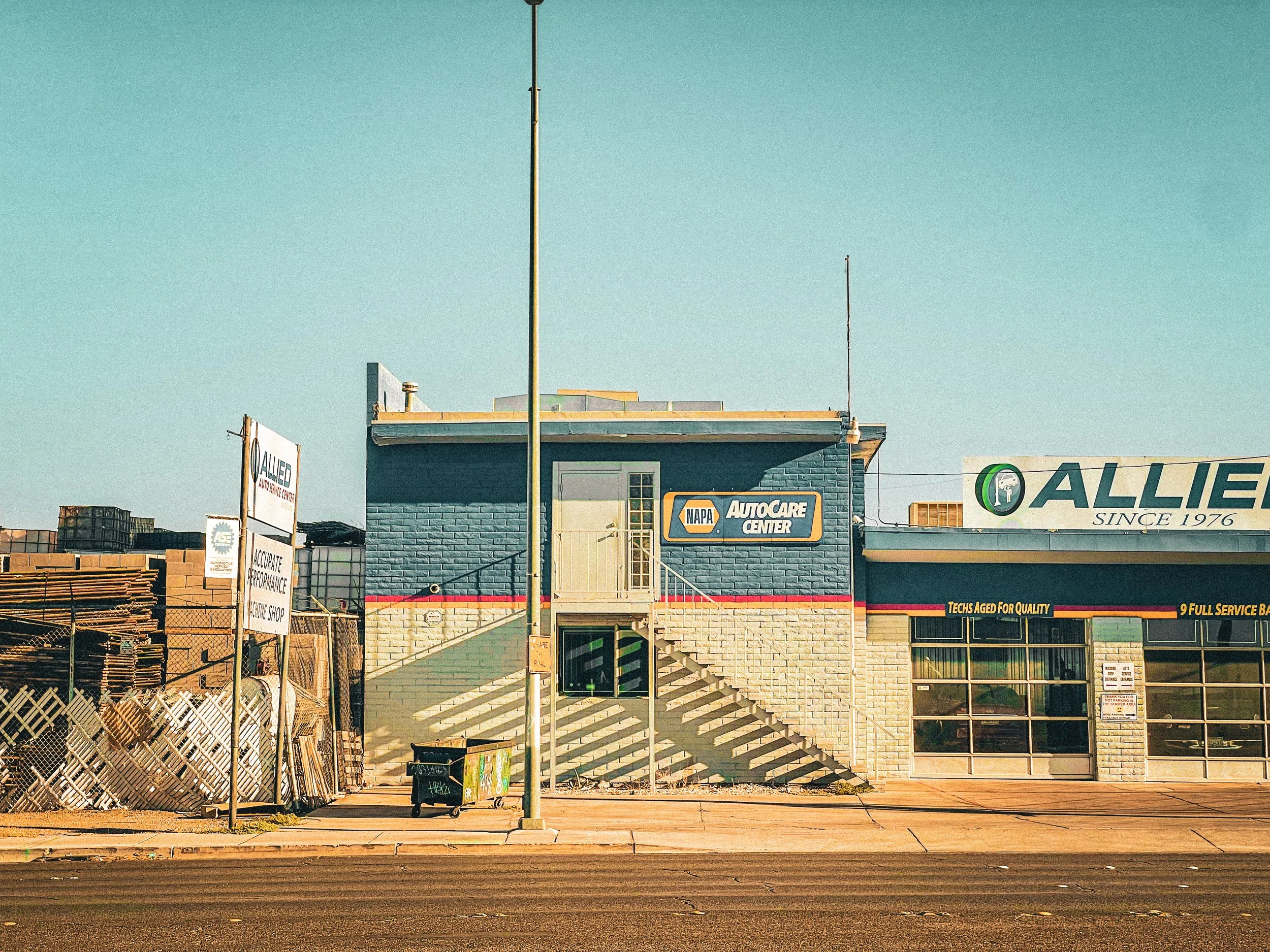 A blue brick auto care shop with stairs leading to a second floor entrance, surrounded by signs and industrial materials, with a clear sky above.