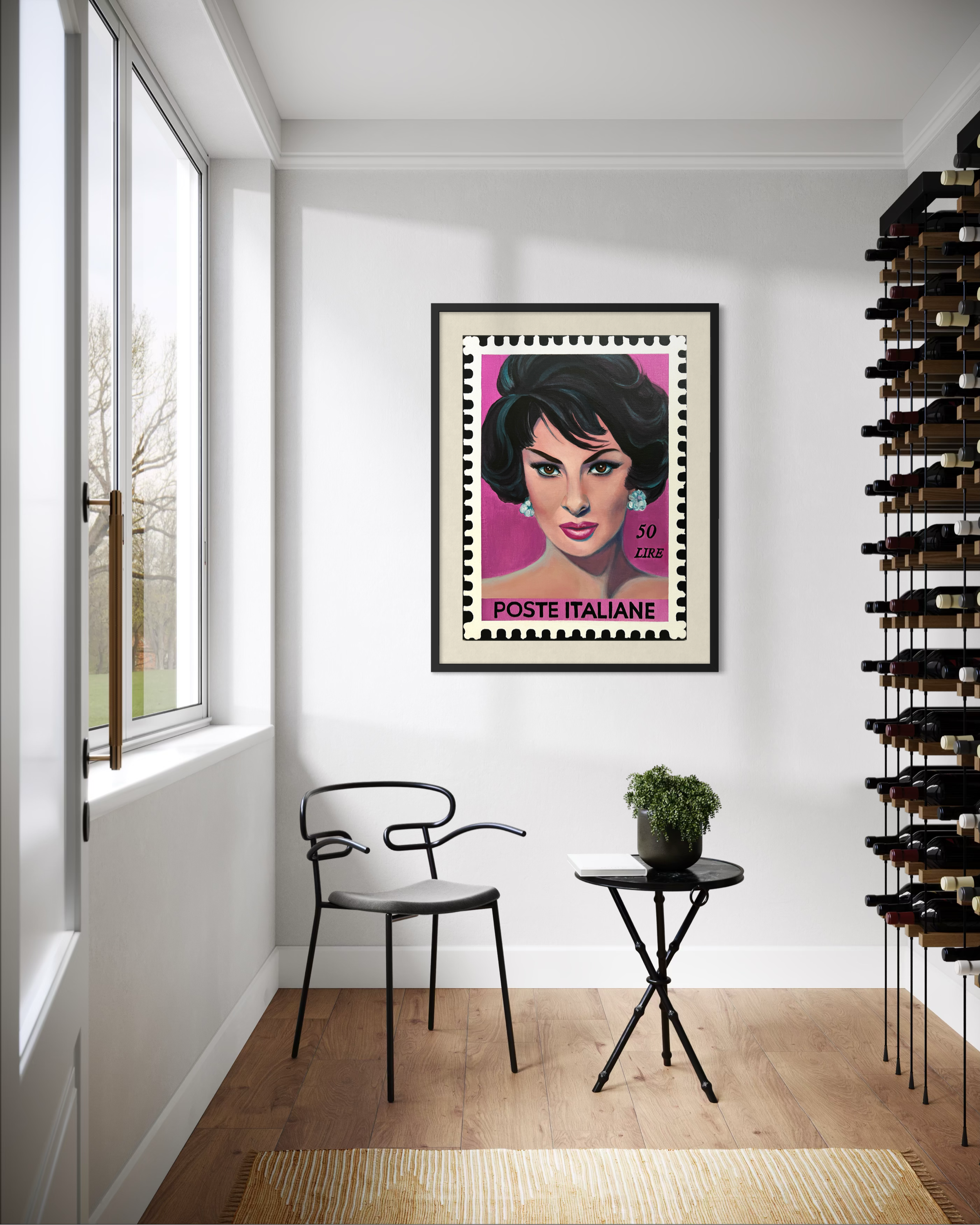 Interior view of a room with a large window on the left and white walls. A framed portrait of a woman with dark hair, earrings, and makeup hangs on the wall. There is a small round table with a book and a potted plant, two black chairs, and a bookshe