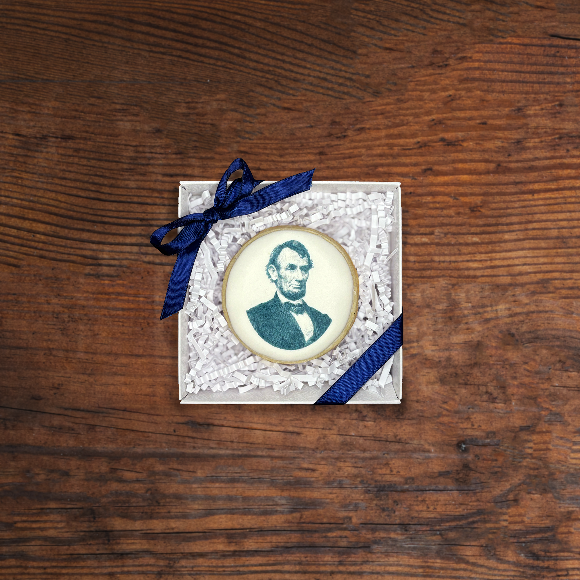 A close-up of a small white gift box with a navy blue ribbon tied in a bow on top, filled with white crinkle paper, containing an oval portrait cookie of Abraham Lincoln on a wooden surface.