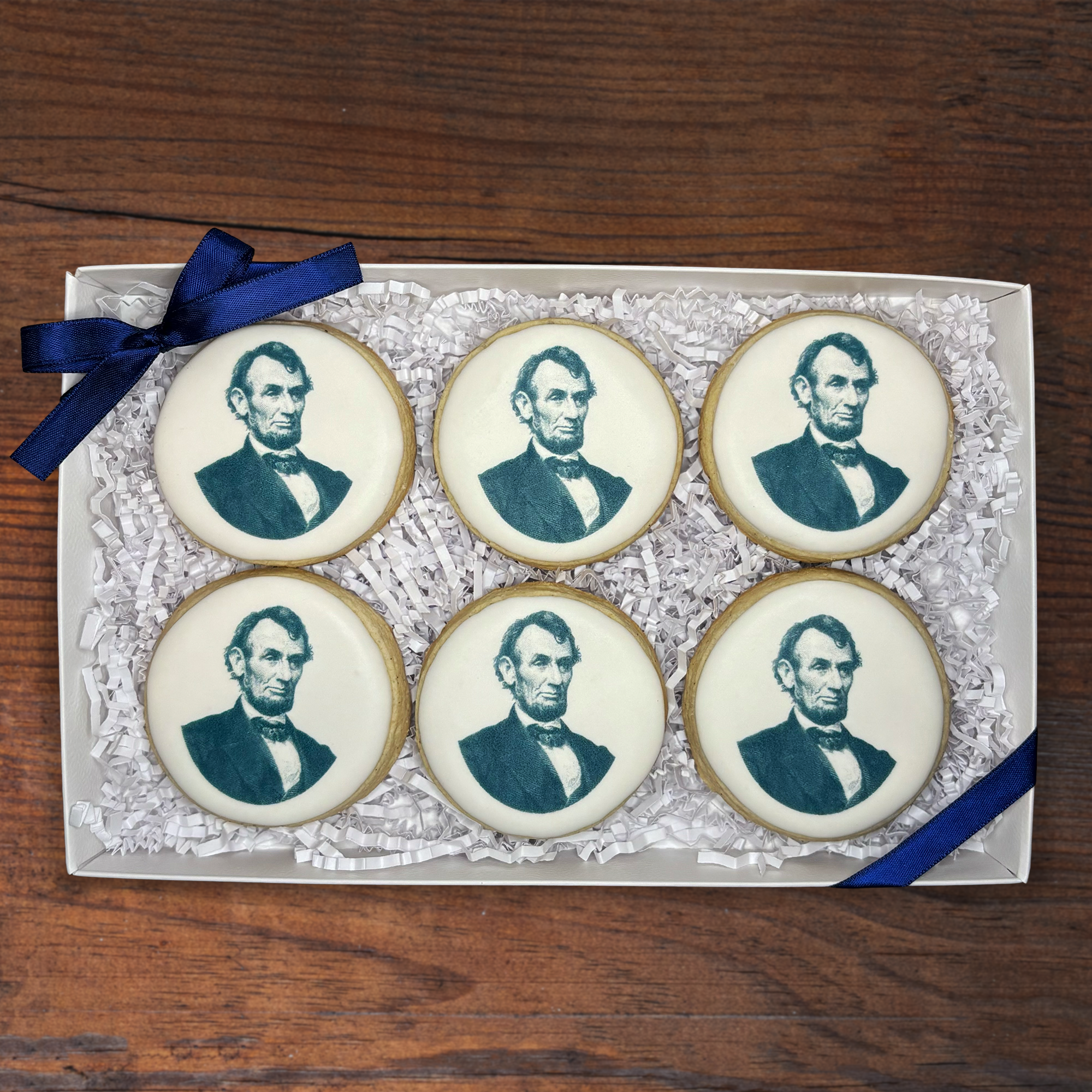 Decorated cookies with Abraham Lincoln's portrait on top, arranged in a box with white paper shreds and a blue ribbon on a wooden surface.
