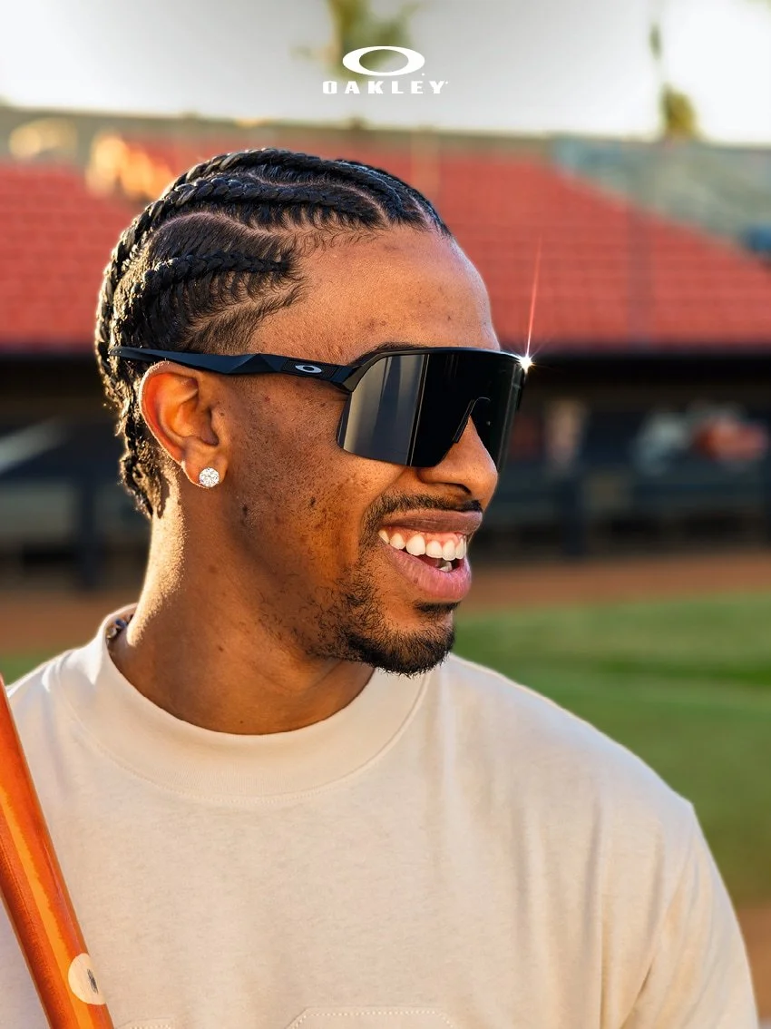 A man with braided hair and diamond earring wearing sunglasses, smiling outdoors. In the background are blurred elements of a residential area. The Oakley logo is visible at the top.