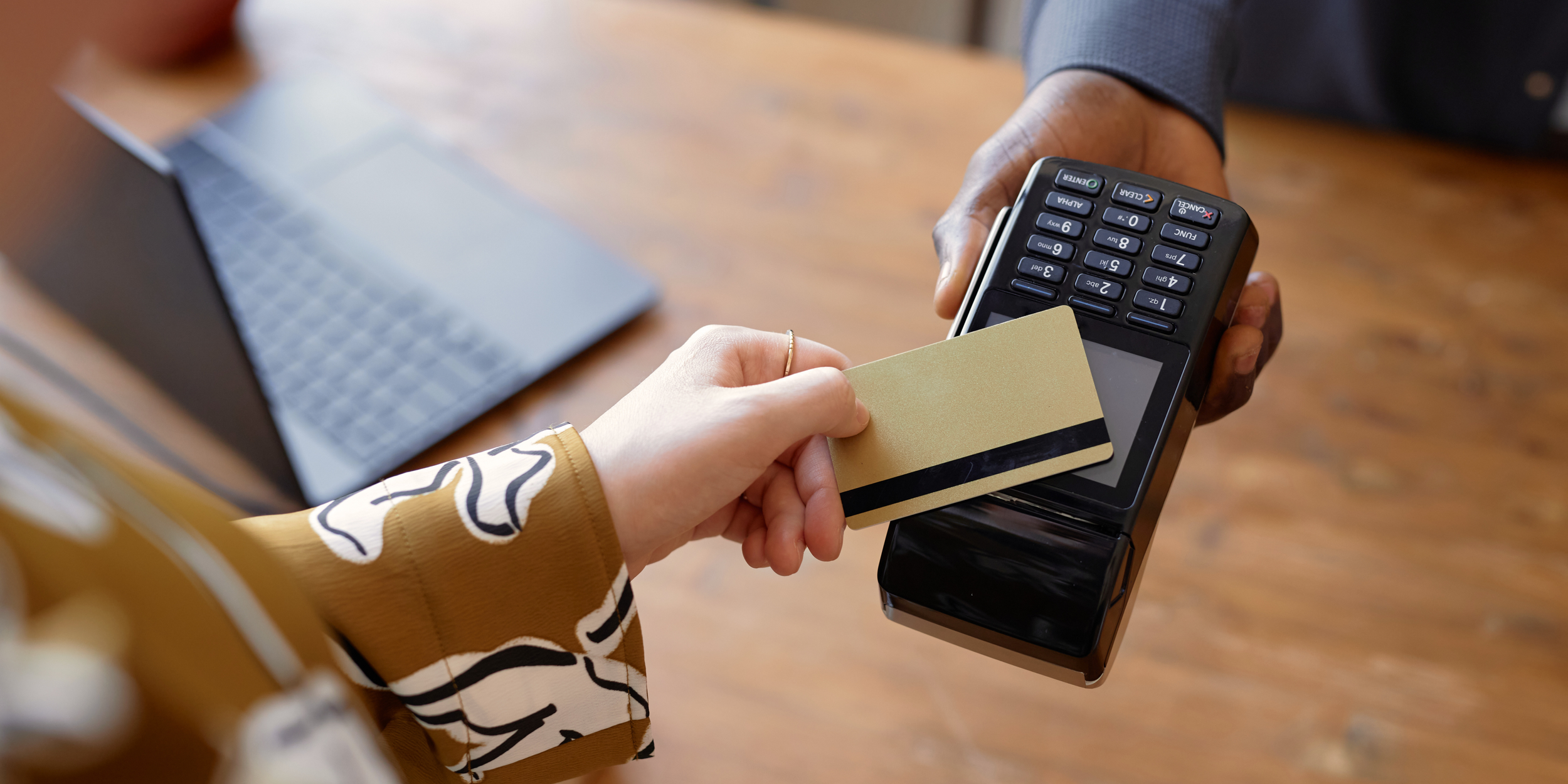 A person is holding a credit card and making a payment on a card terminal, with a laptop in the background on a wooden table.