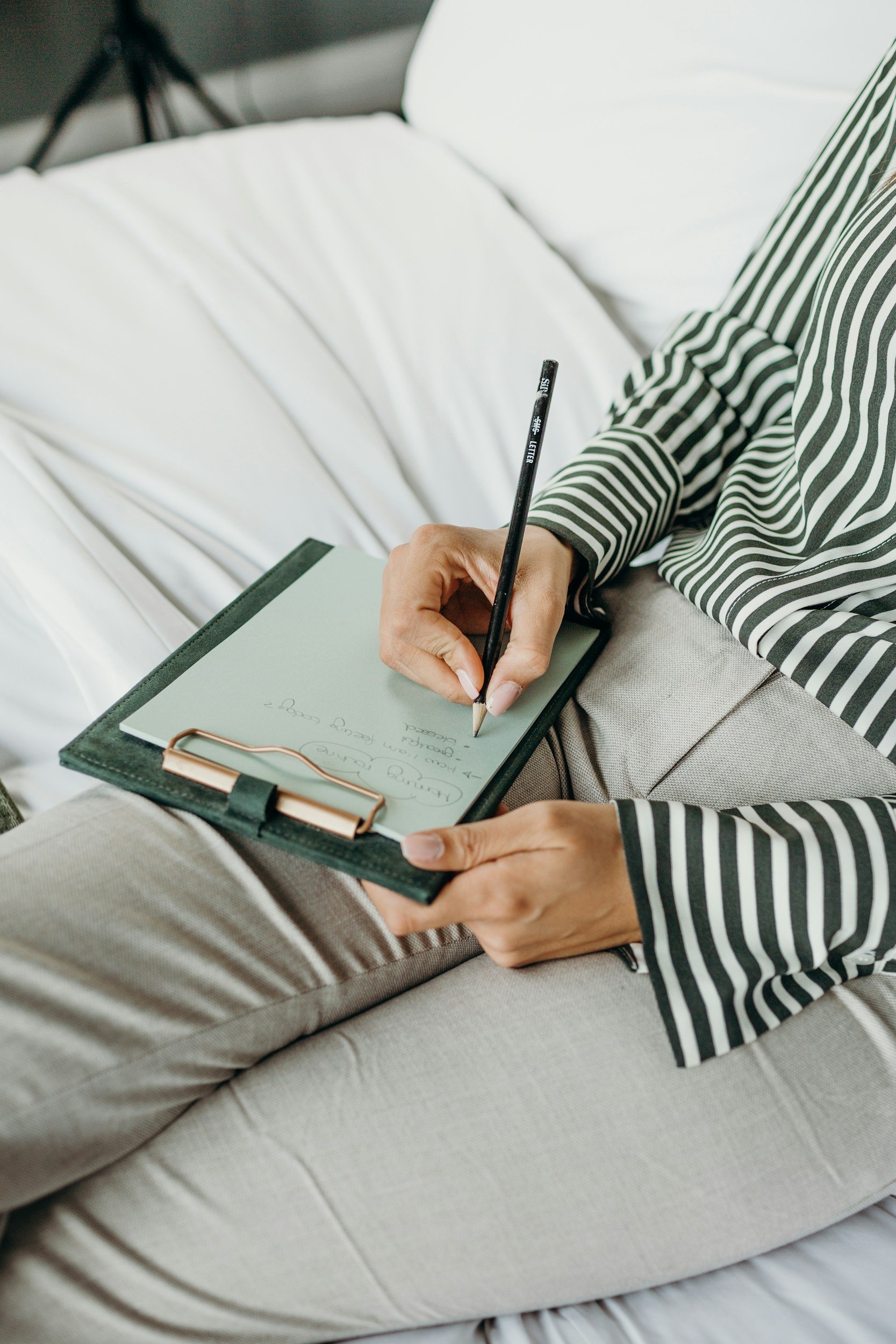 Person sitting on a beige couch writing in a notebook with a black pen, wearing a black and white striped shirt.