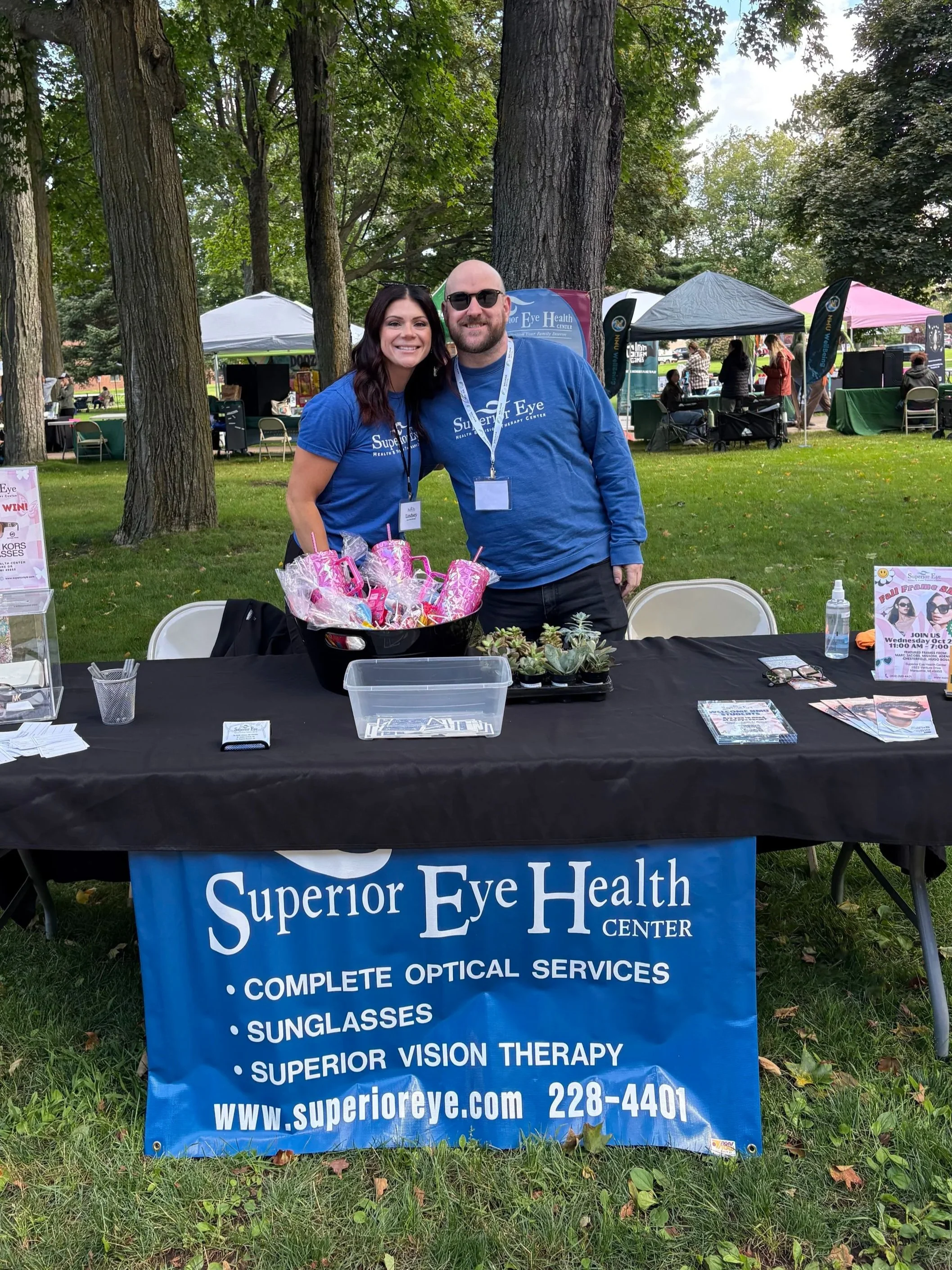 Two smiling people wearing blue shirts with the words 'Superior Eye' are standing behind a table at an outdoor event. The table displays various promotional items, and a large blue banner with white text advertises their eye care services. They are i