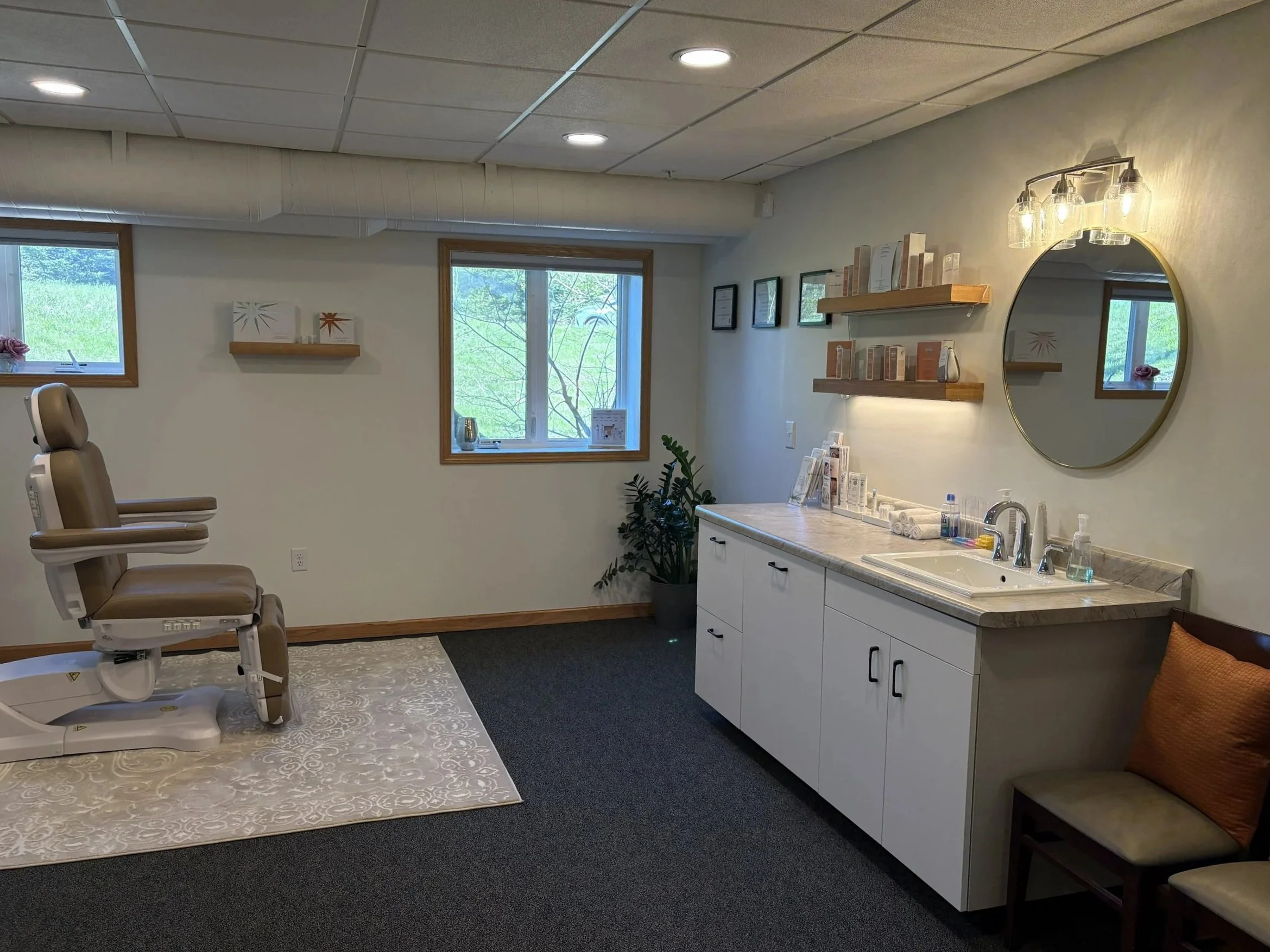 Medical examination room with an exam chair, windows with greenery outside, shelves with books or pamphlets, a sink, and a round mirror with light fixtures.